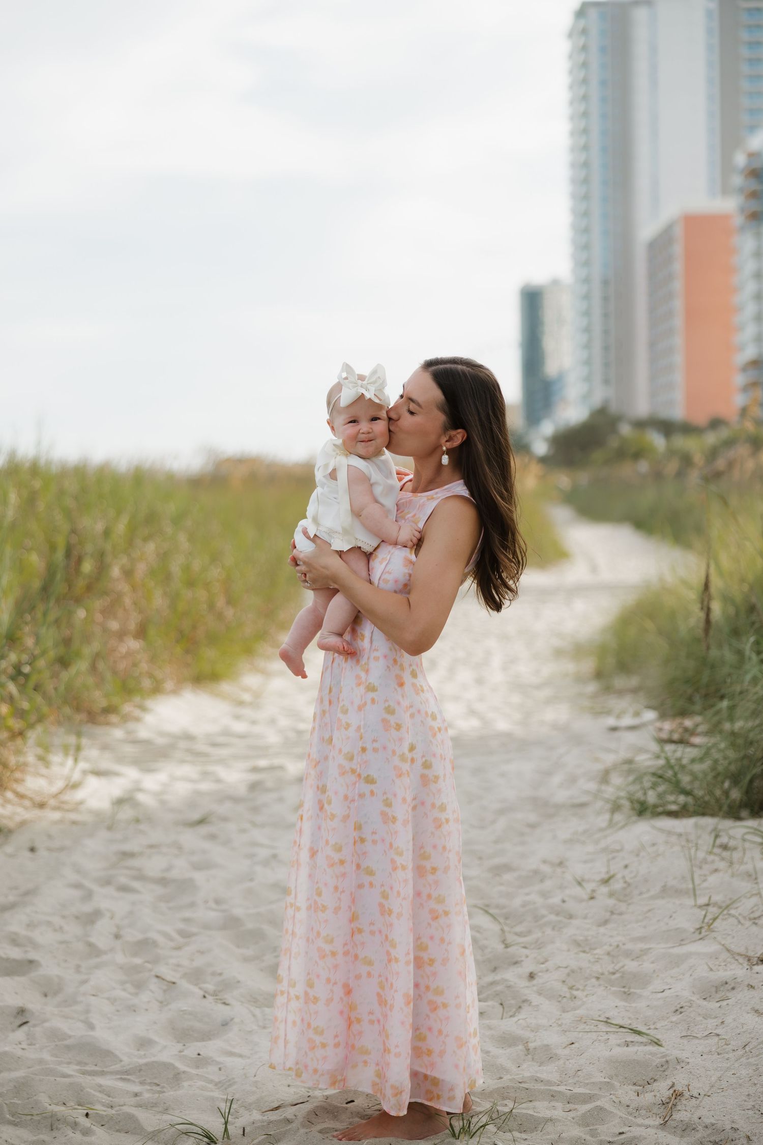 Mother in floral maxi dress holds baby girl in white outfit on sandy beach path near coastal buildings.