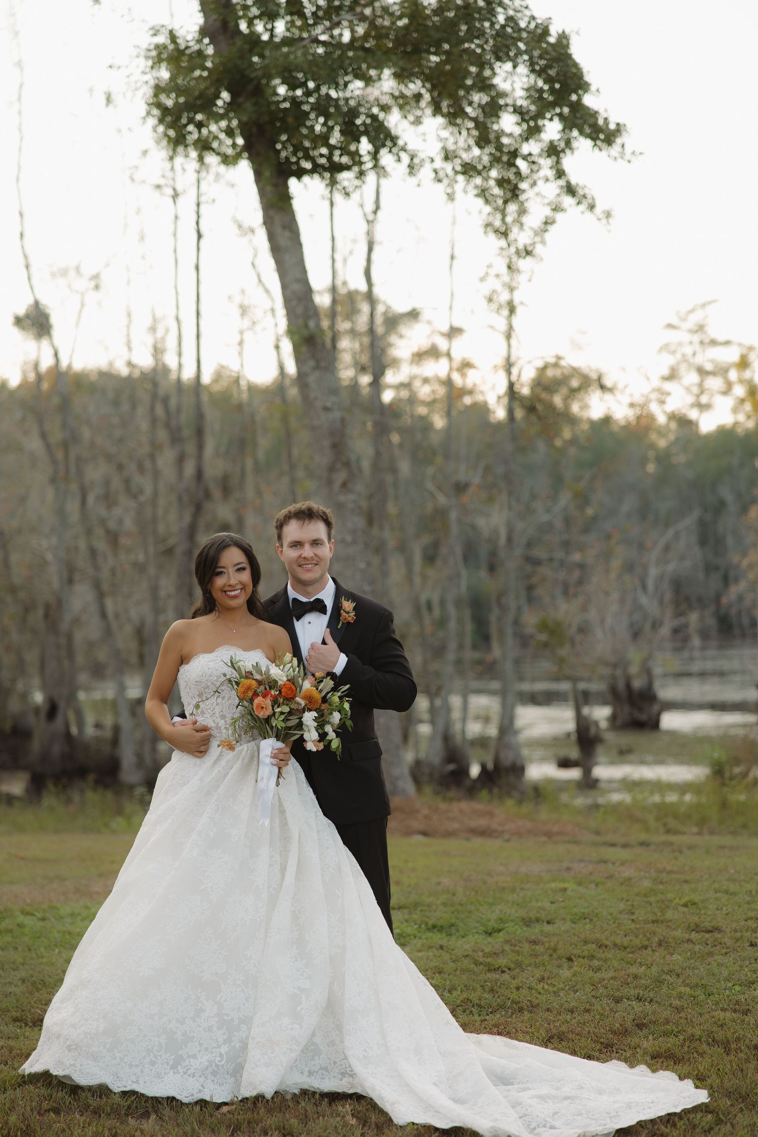 Bride in a strapless lace wedding gown and groom in a black tuxedo hold an orange floral bouquet on a grassy lawn near a swamp.