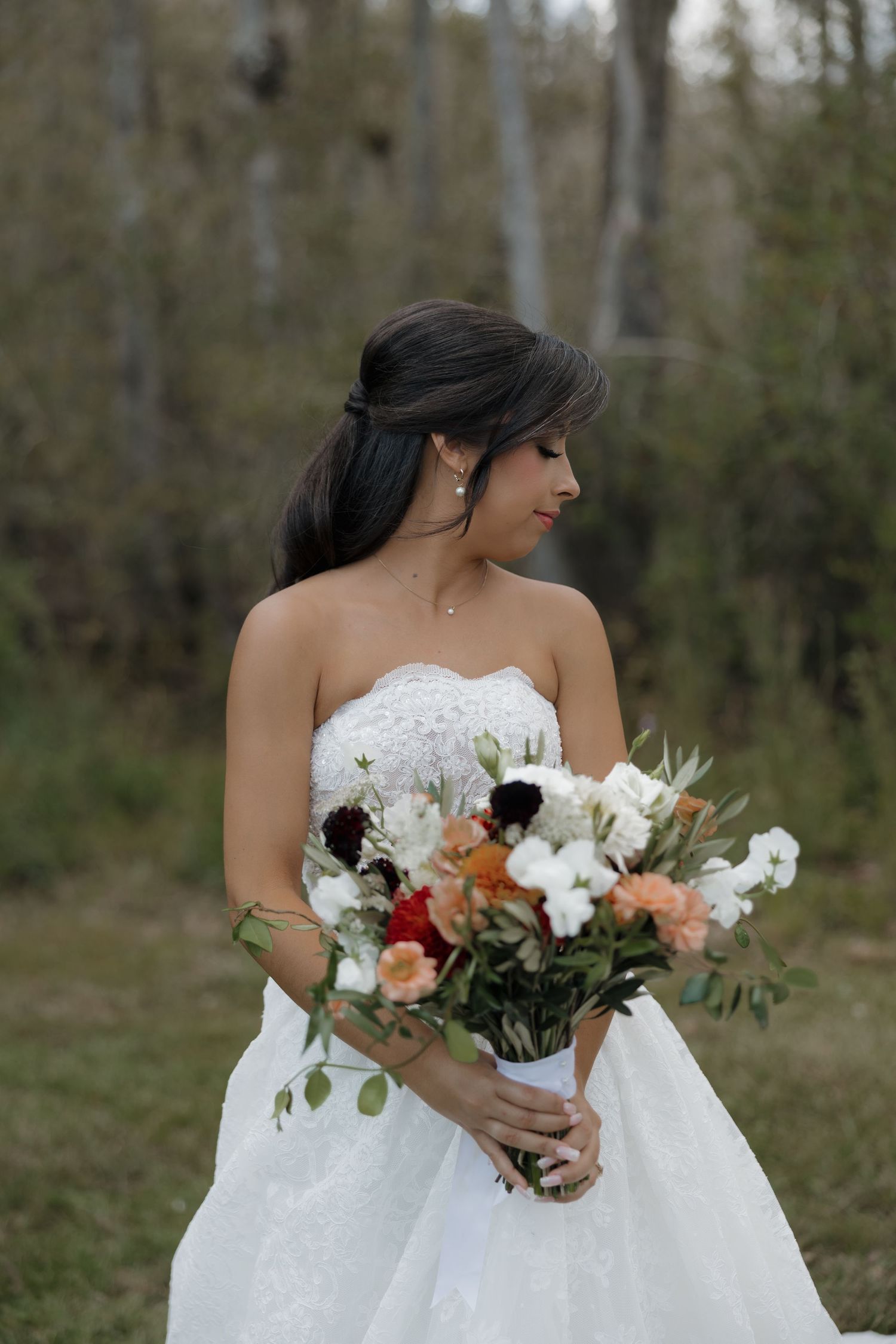Bride in a strapless white lace gown holds an orange, white and burgundy bouquet while standing in a wooded outdoor setting.