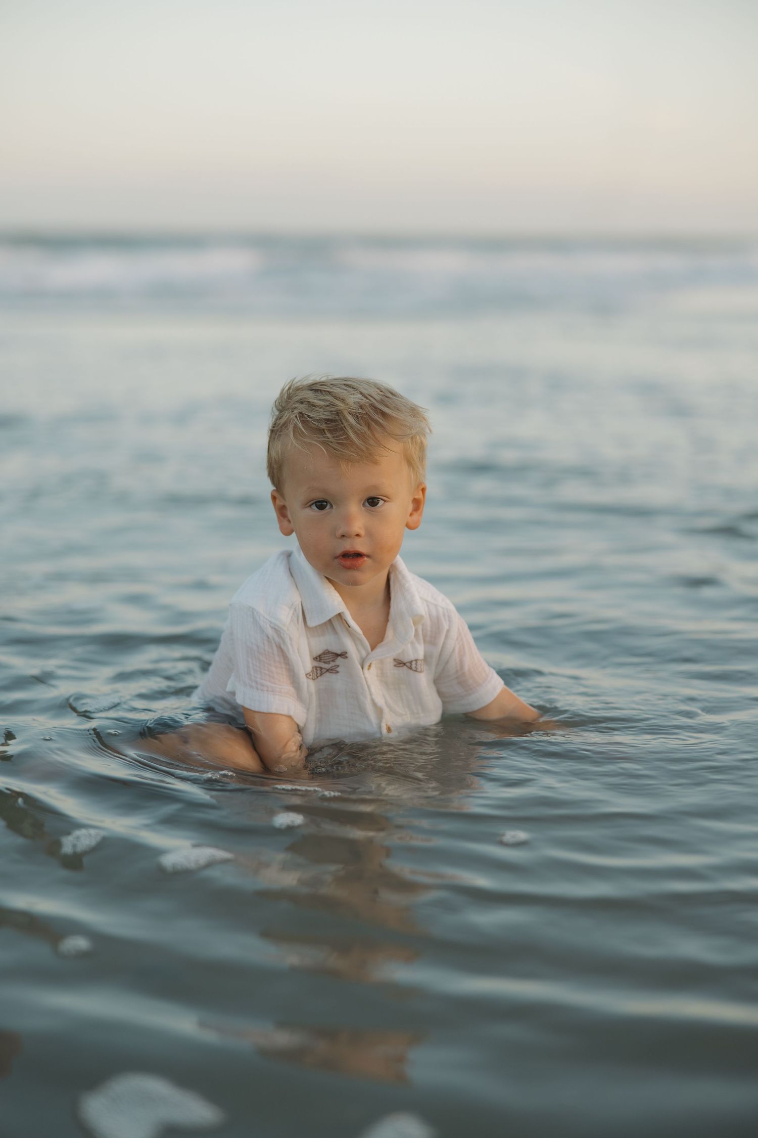 Young blonde toddler boy in white shirt wading in ocean waves at beach during sunset.