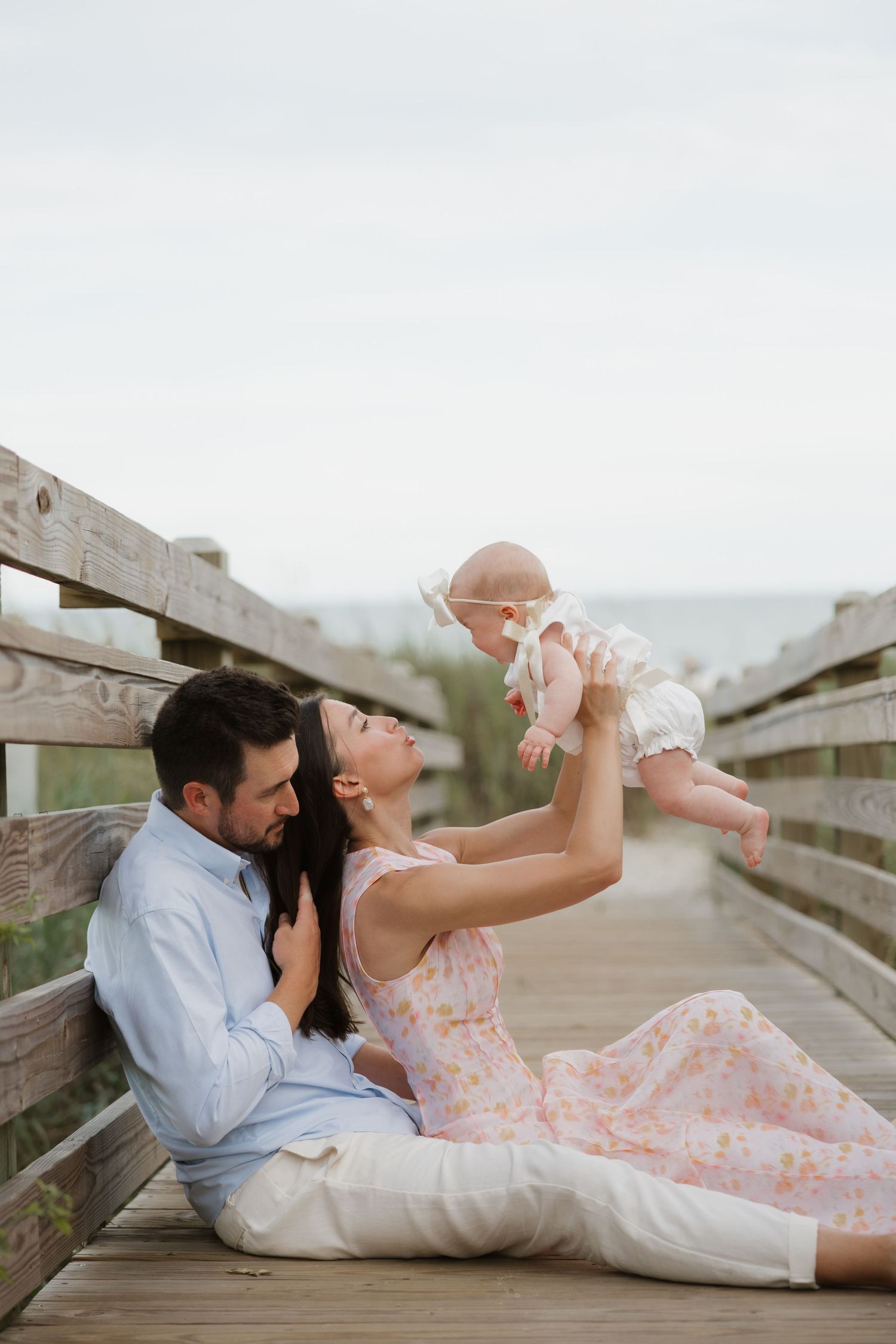 A couple sits on a beach boardwalk, smiling as the mother lifts their baby girl in the air.