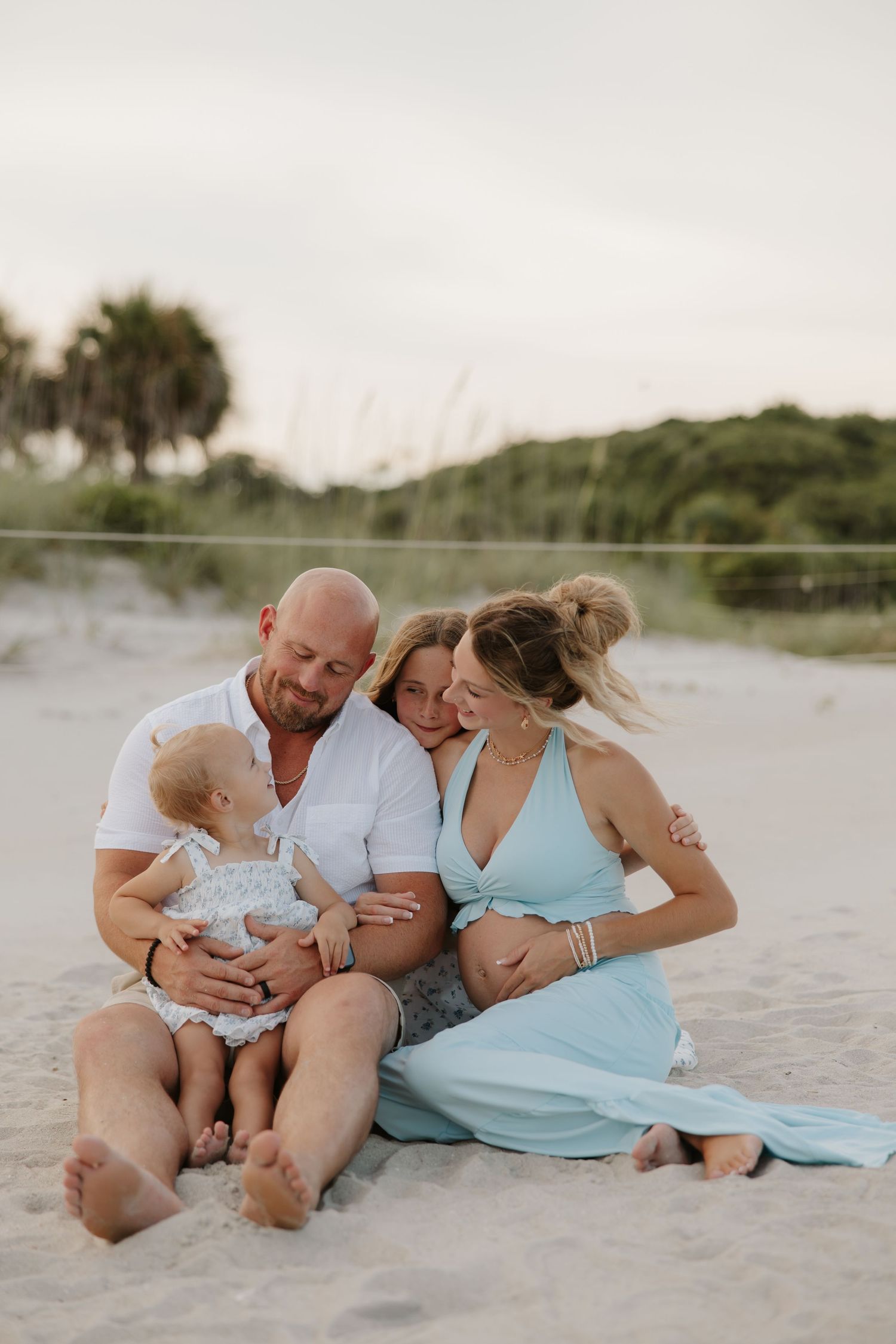 A happy family sits on a sandy beach, with a bald man, pregnant woman in teal dress, and baby girl.