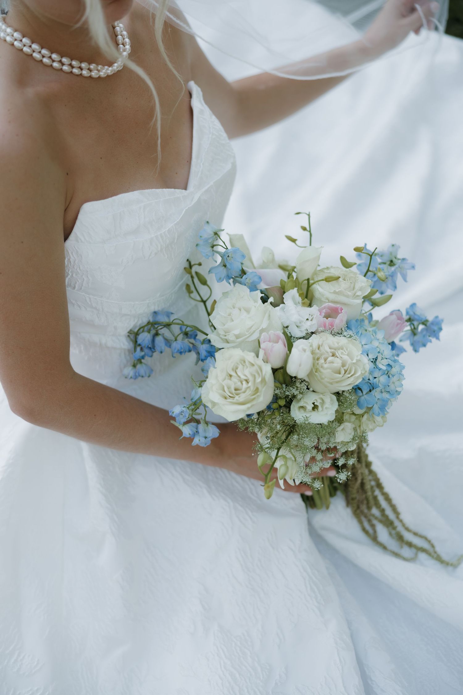Bride in white strapless gown holding a bouquet of white roses and blue flowers, wearing a pearl necklace.