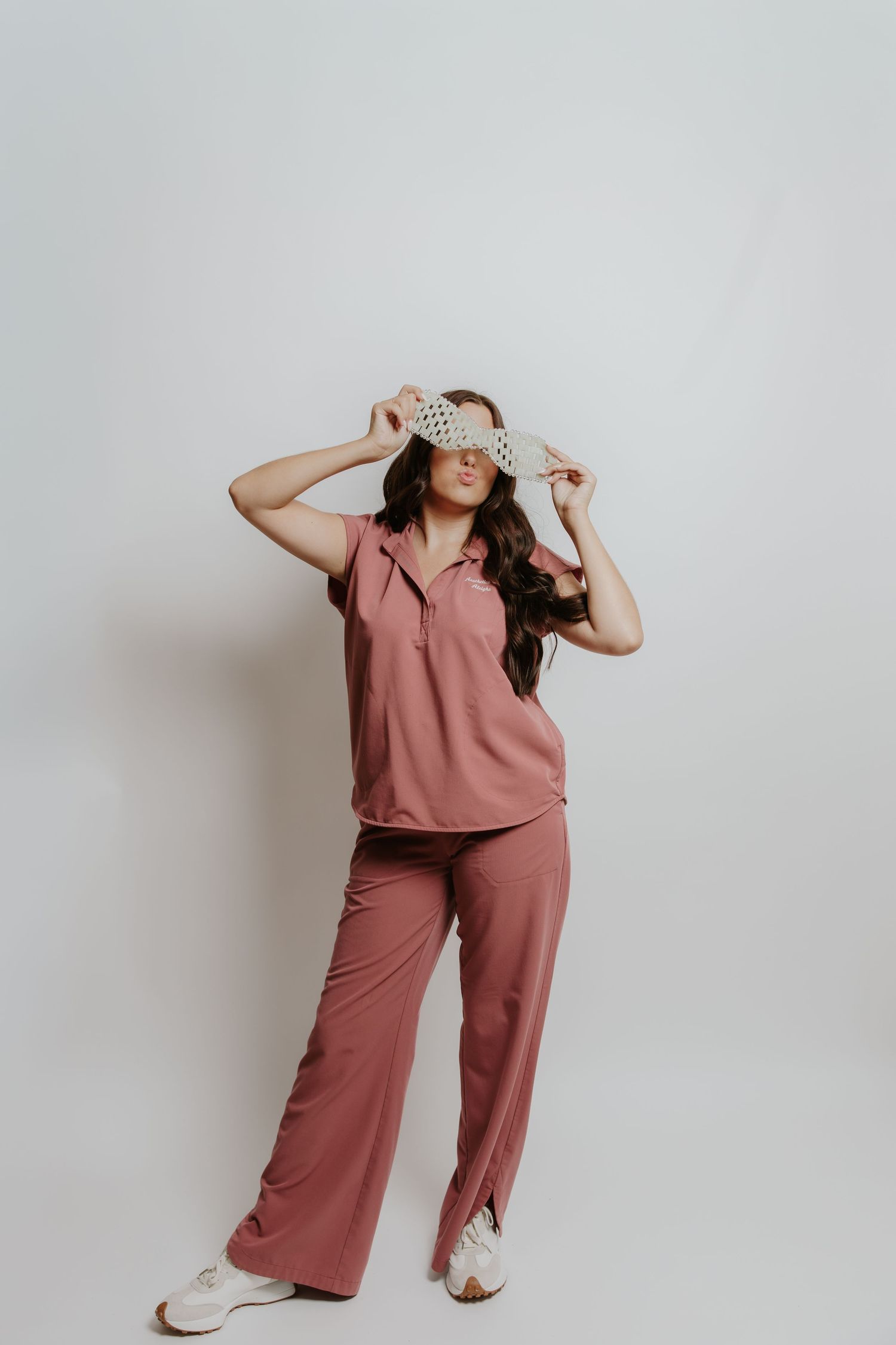 Woman in pink matching pajama set holding a sleep mask over her eyes on white background.