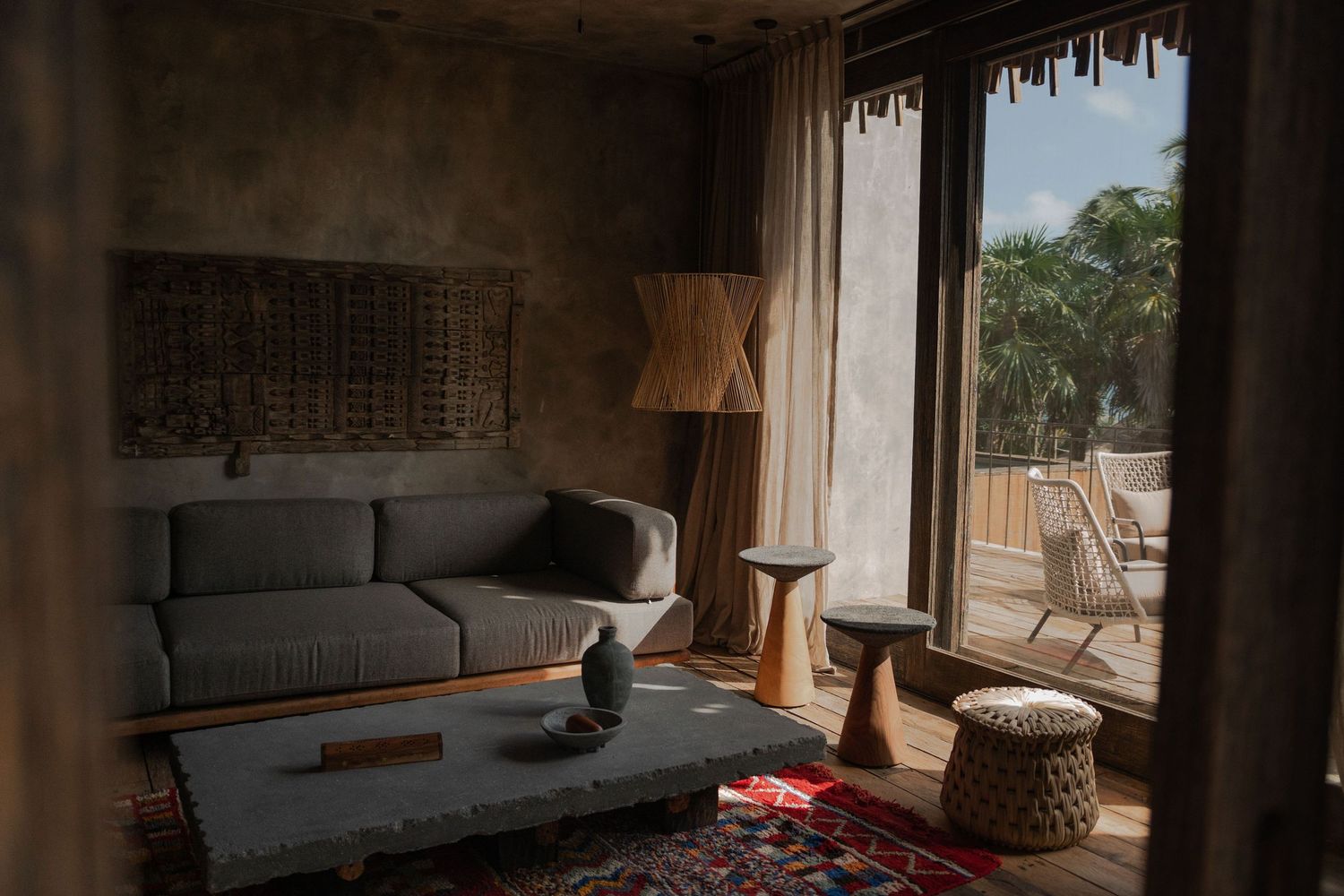 A cozy modern living room features a gray sofa, woven pendant light, and large windows overlooking tropical greenery.