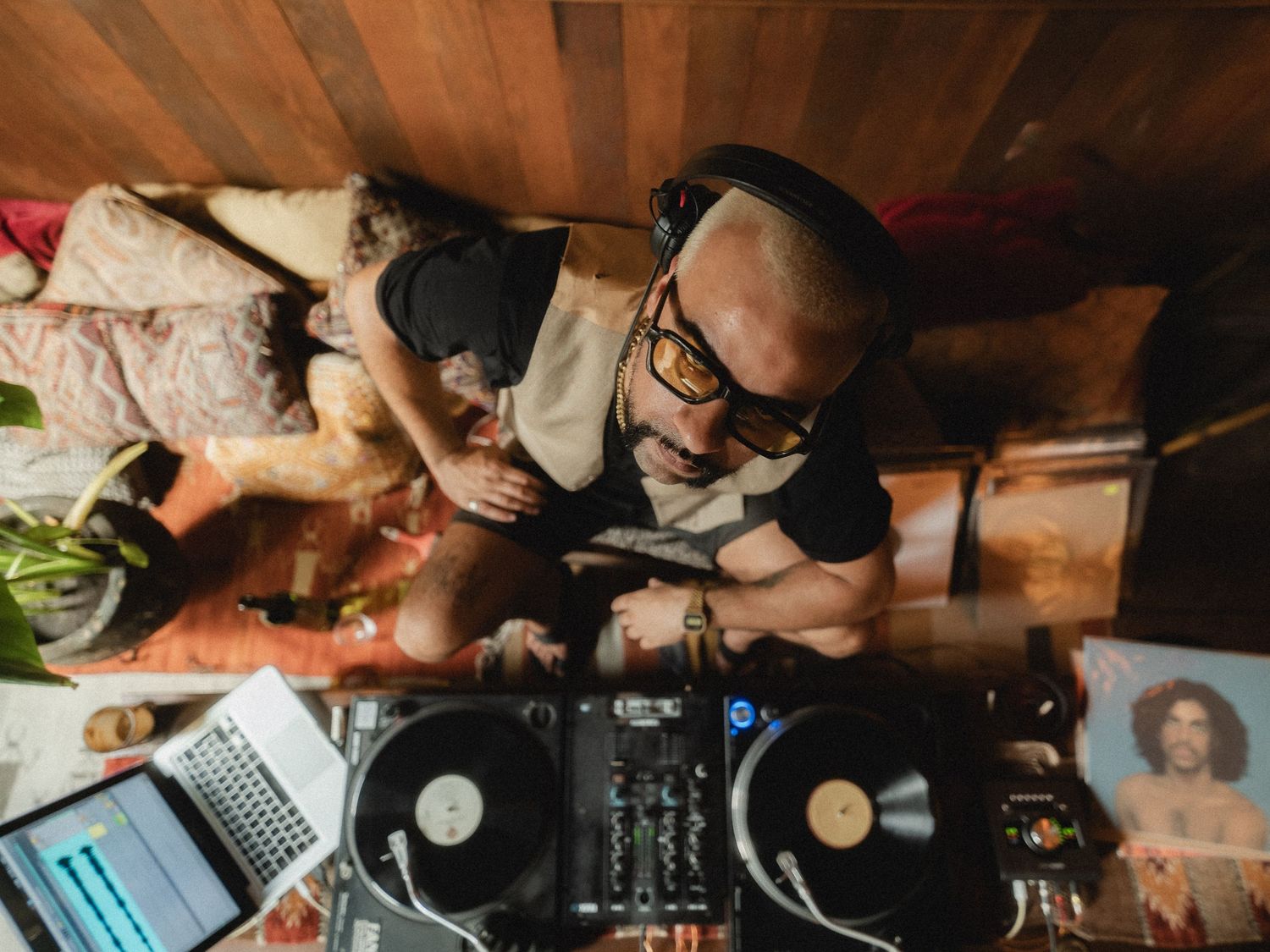 Overhead view of a DJ with headphones and glasses spinning vinyl records on turntables in a cozy home studio.
