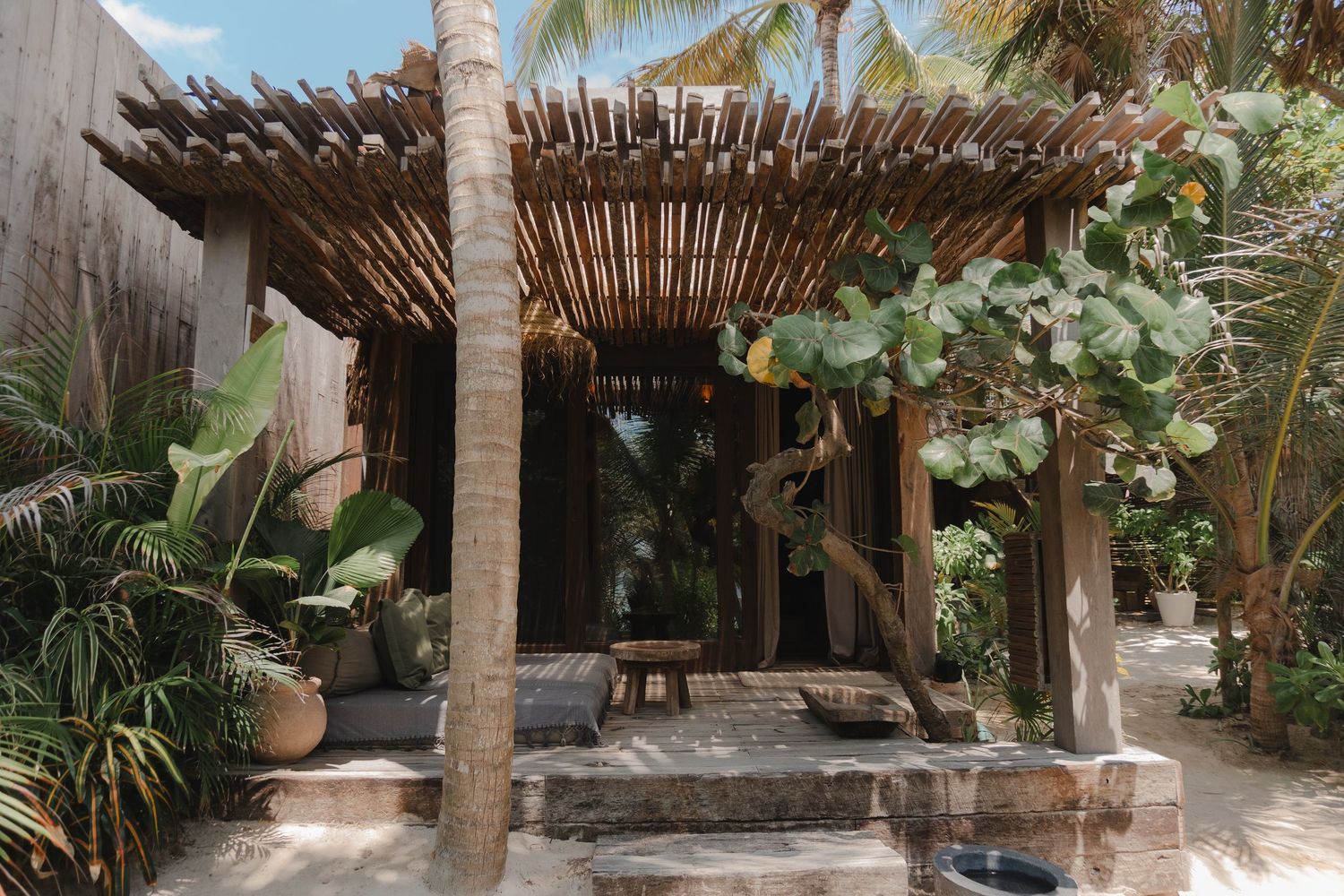Rustic wooden pergola with palm trees creates a tropical beach house patio atmosphere in a coastal setting.