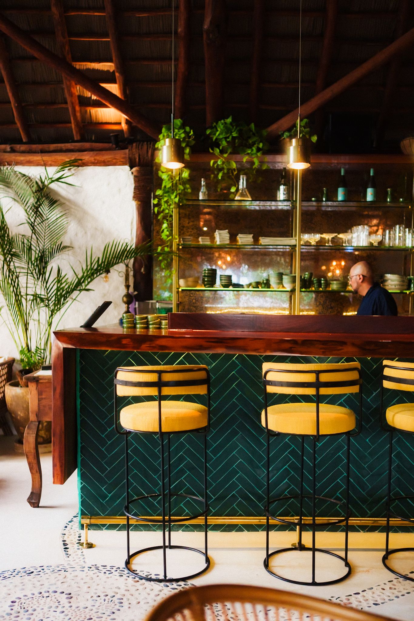Modern bar interior with green chevron pattern and yellow bar stools alongside tropical plants and warm lighting.