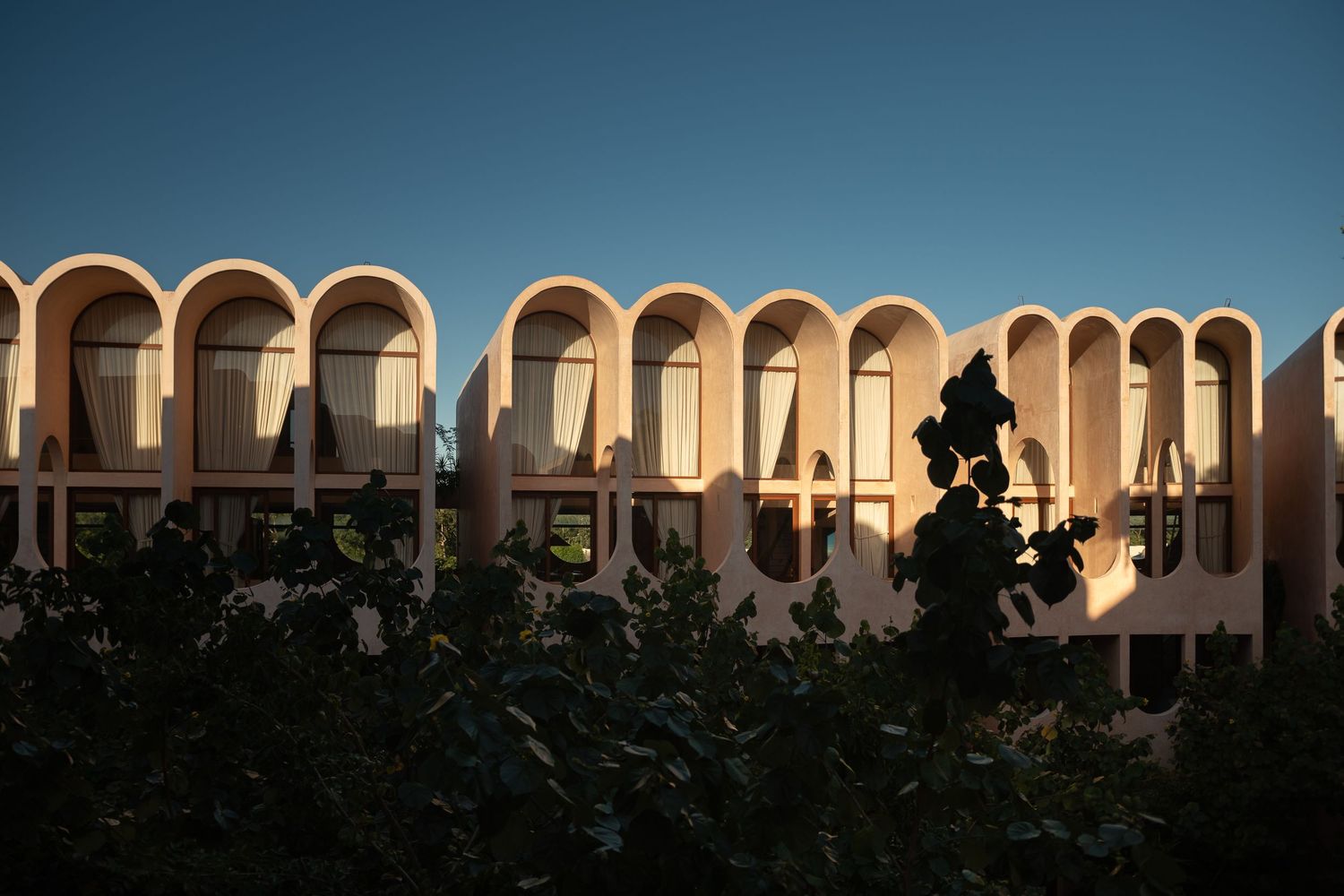 Arched facade of a modernist building bathed in golden sunset light, with silhouetted foliage in the foreground.