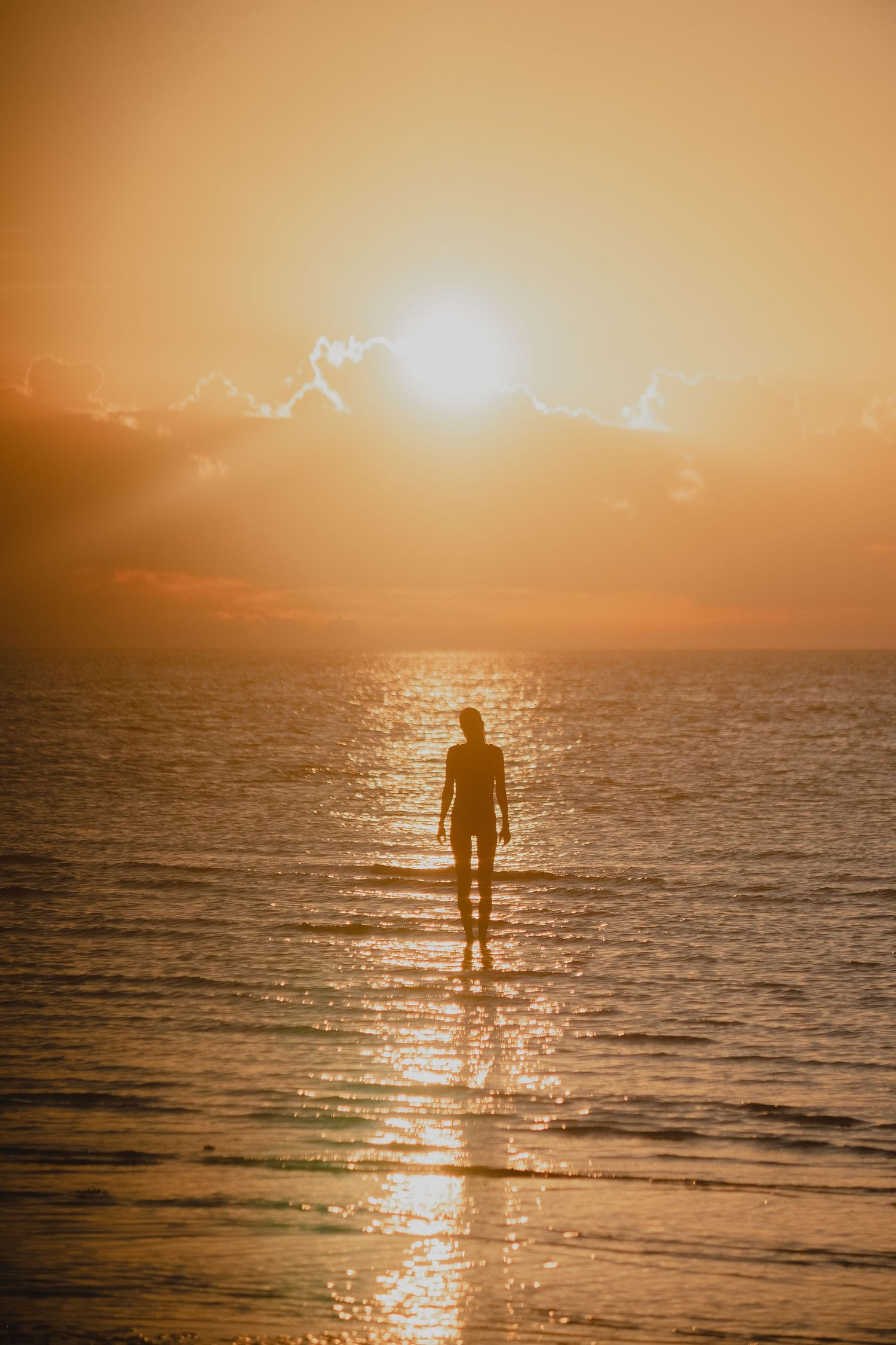 A silhouetted figure stands in shallow ocean water during golden hour as sunlight sparkles on the waves at sunset.