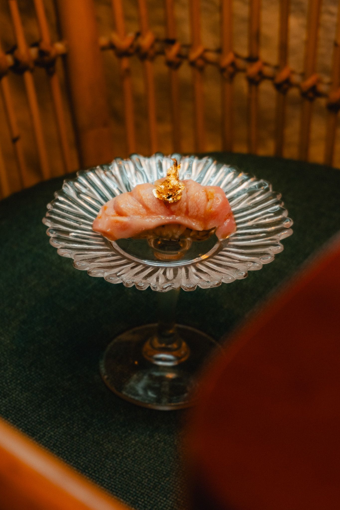 A piece of fresh salmon sashimi garnished with gold flakes served on a crystal glass plate in dim lighting.
