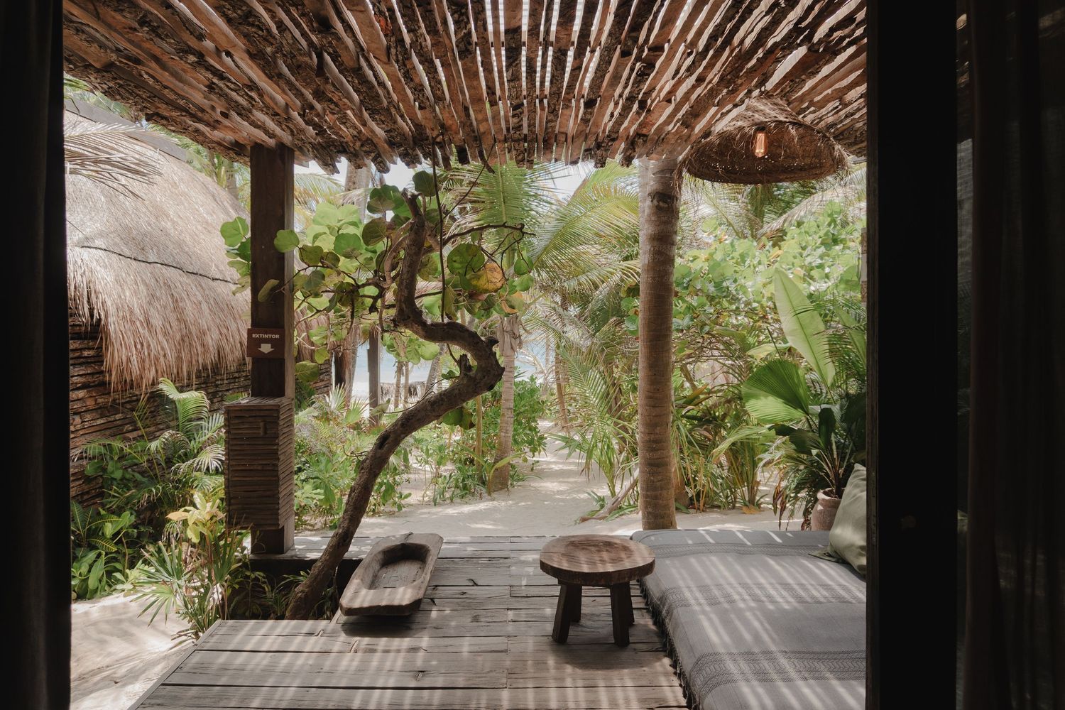 A wooden deck with thatched roof pergola extends into a lush tropical garden setting with stone pathways.