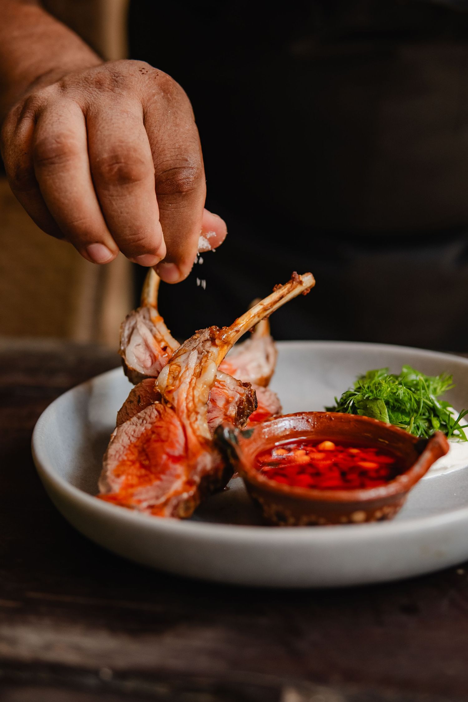 Hands holding grilled shrimp with red dipping sauce on white plate.