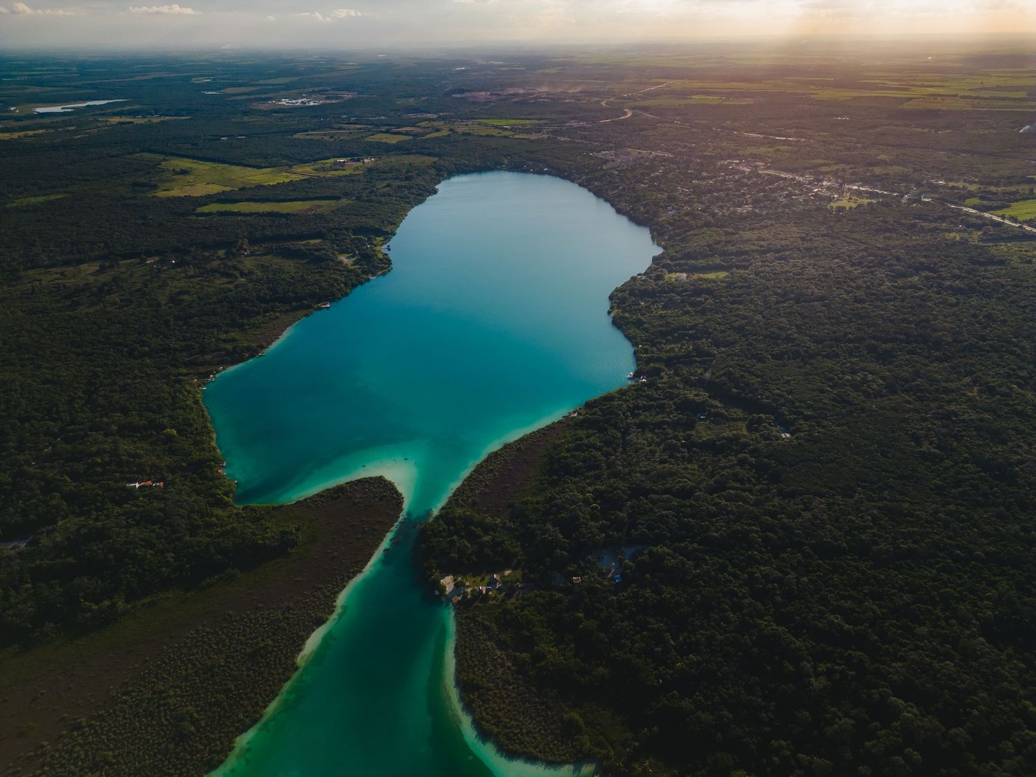 Aerial view of a turquoise lake with narrow channel surrounded by dense forest during sunset.