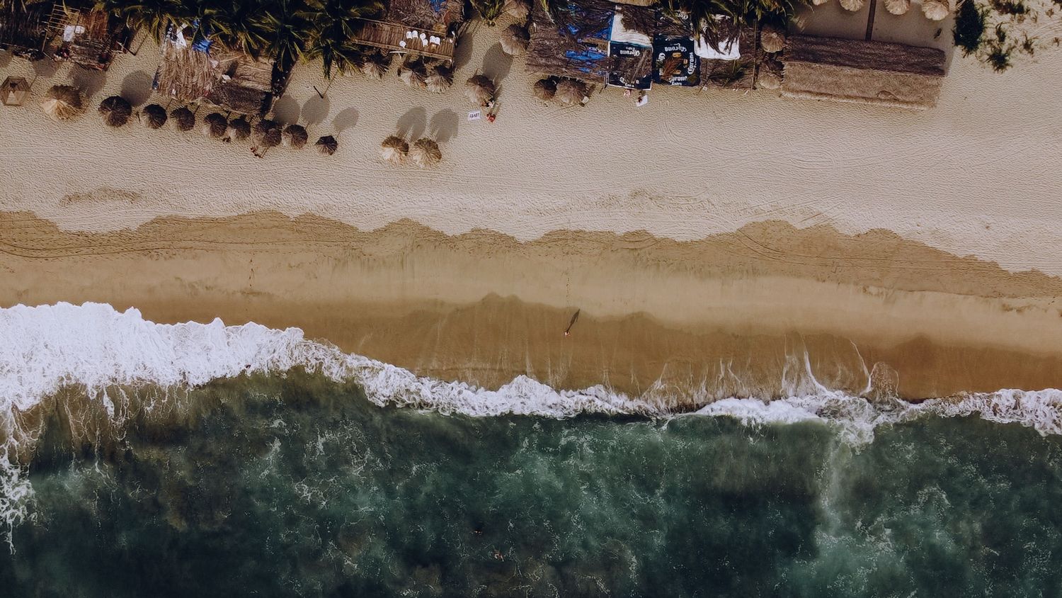 Aerial drone view of a sandy beach coastline with turquoise ocean waters and waves breaking along the shore.