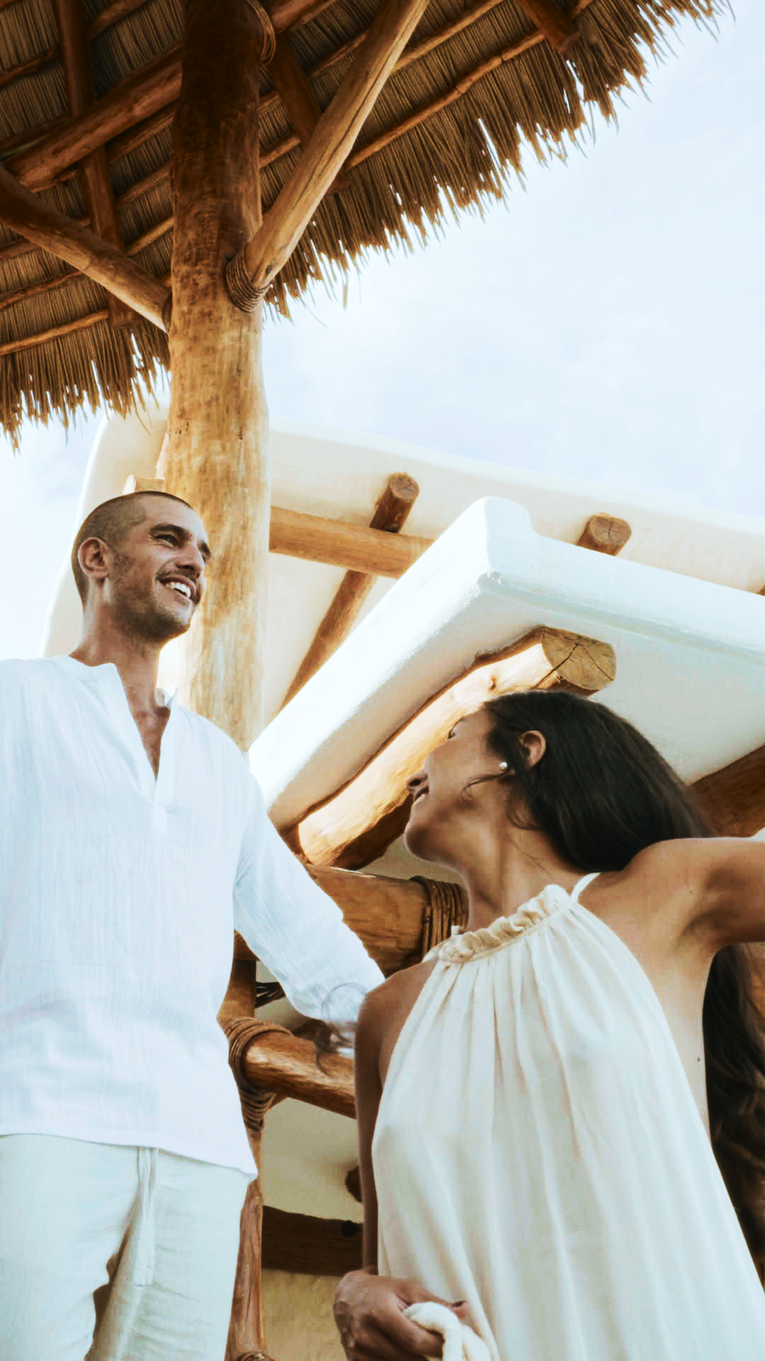 A couple in white outfits share a romantic moment under a thatched roof pergola at a tropical resort.