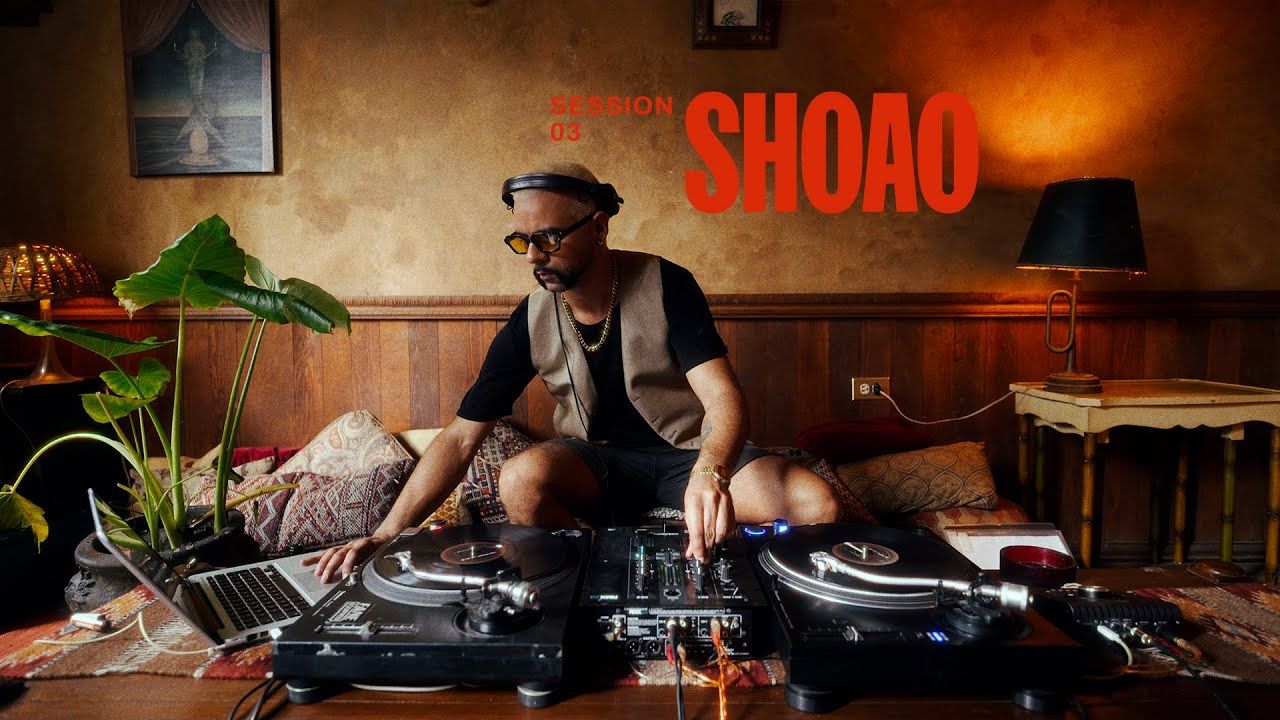 A DJ performs with vinyl turntables and mixing equipment in a dimly lit nightclub setting.
