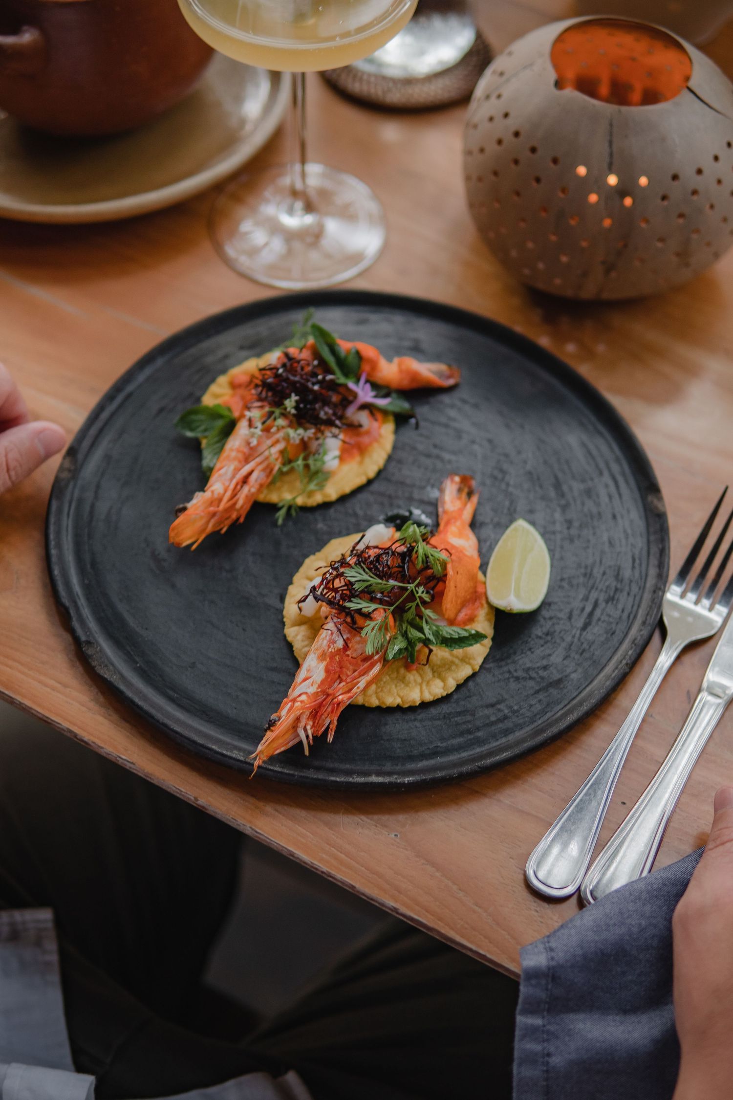 Grilled shrimp appetizers served on black plates with lime garnish and fresh herbs on a wooden dining table.