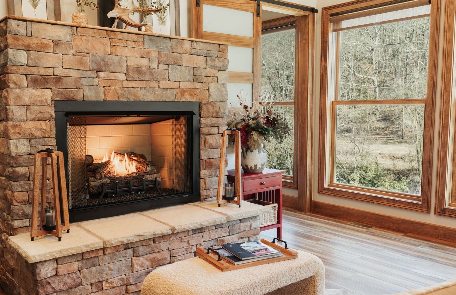 Warm stone fireplace with a books in front of it. Large windows showing forest.