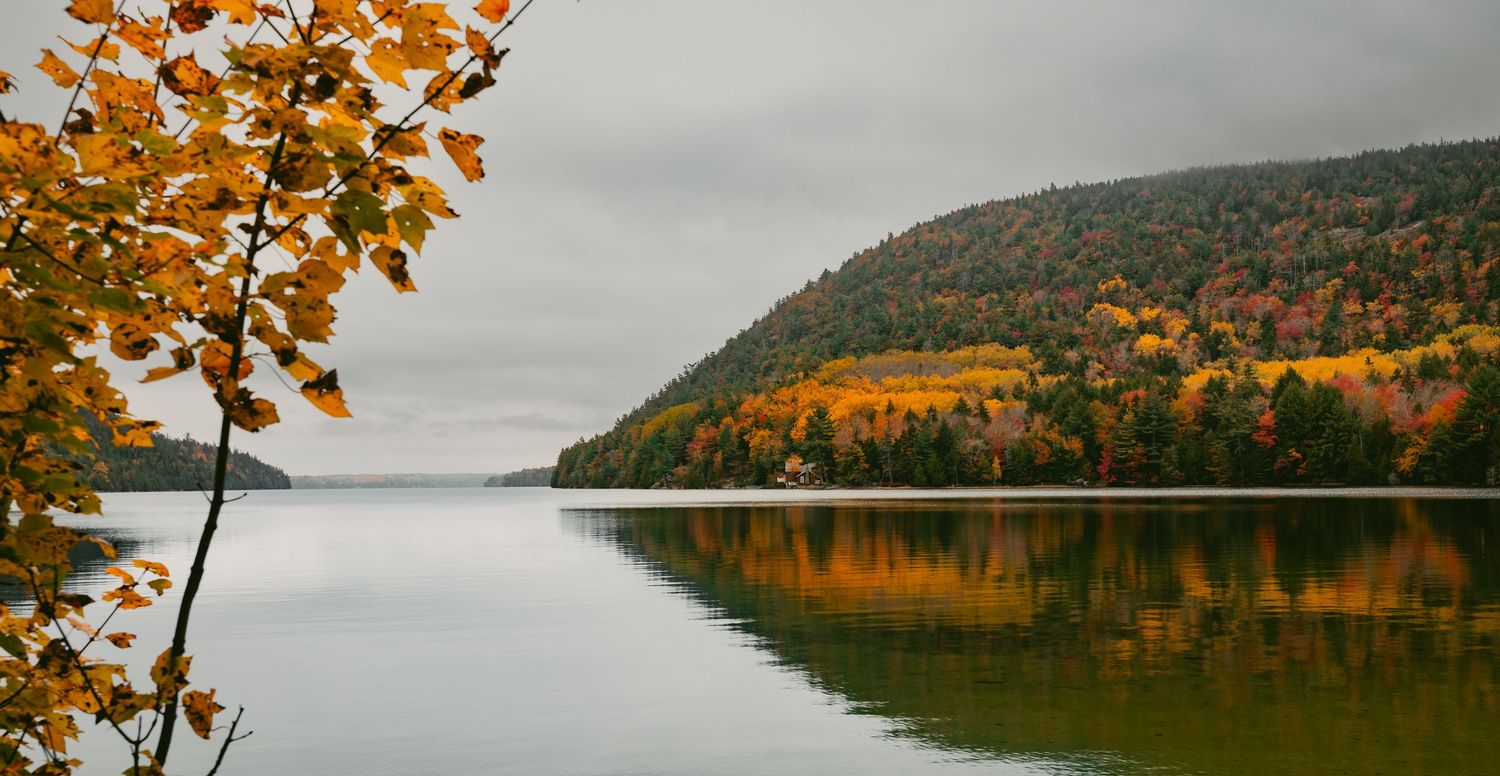 A lake surrounded by colorful trees in the fall with a small brown cabin off in the distance.