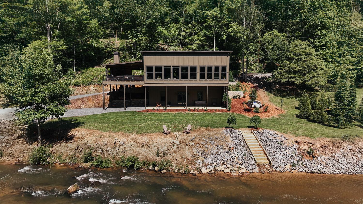 Straight on view of a cabin overlooking a river with stone stairs leading down to the water. The cabin is surrounded by trees.