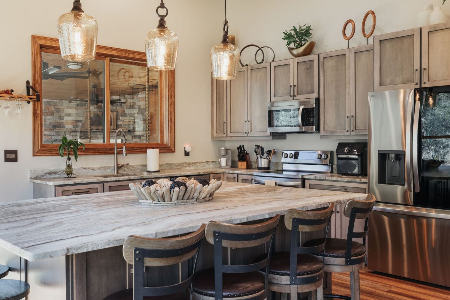A kitchen with gray cabinets and natural stone countertops. Three pendant lights dangle from the ceiling. A large window with wood trim.