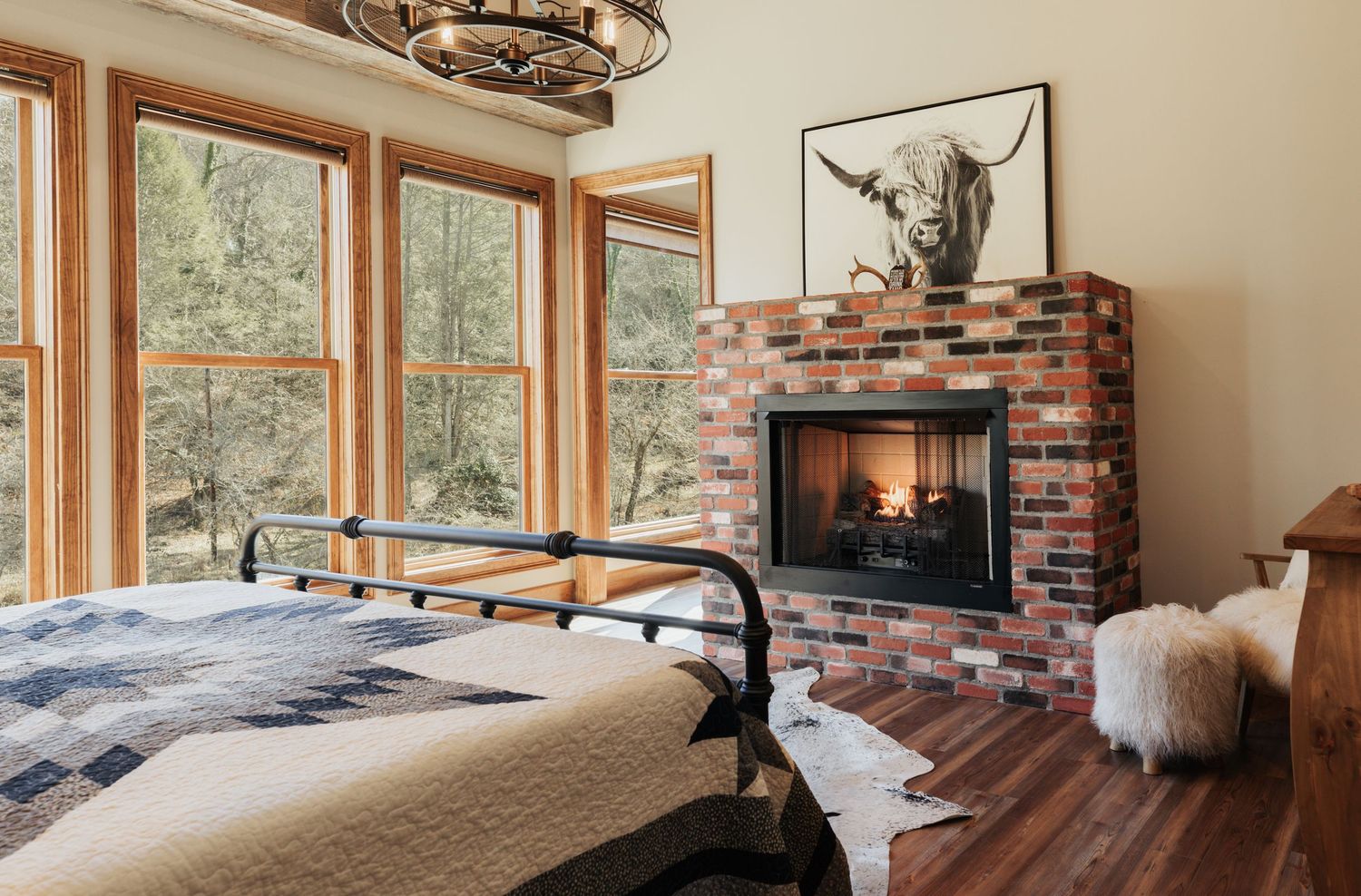 A bedroom with large windows overlooking wilderness, with a brick fireplace, cowhide carpet, and a small furry stool.
