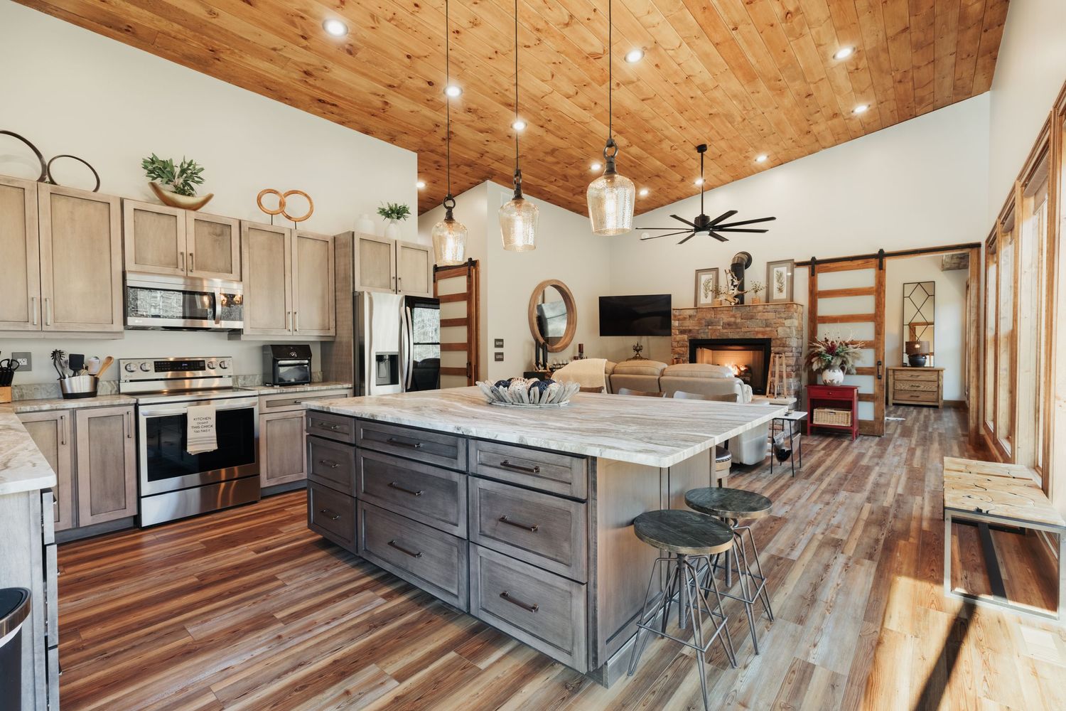 Open floor plan showing a kitchen island with barstools on the end. Wood ceilings with three pendent lights and a large ceiling fan.