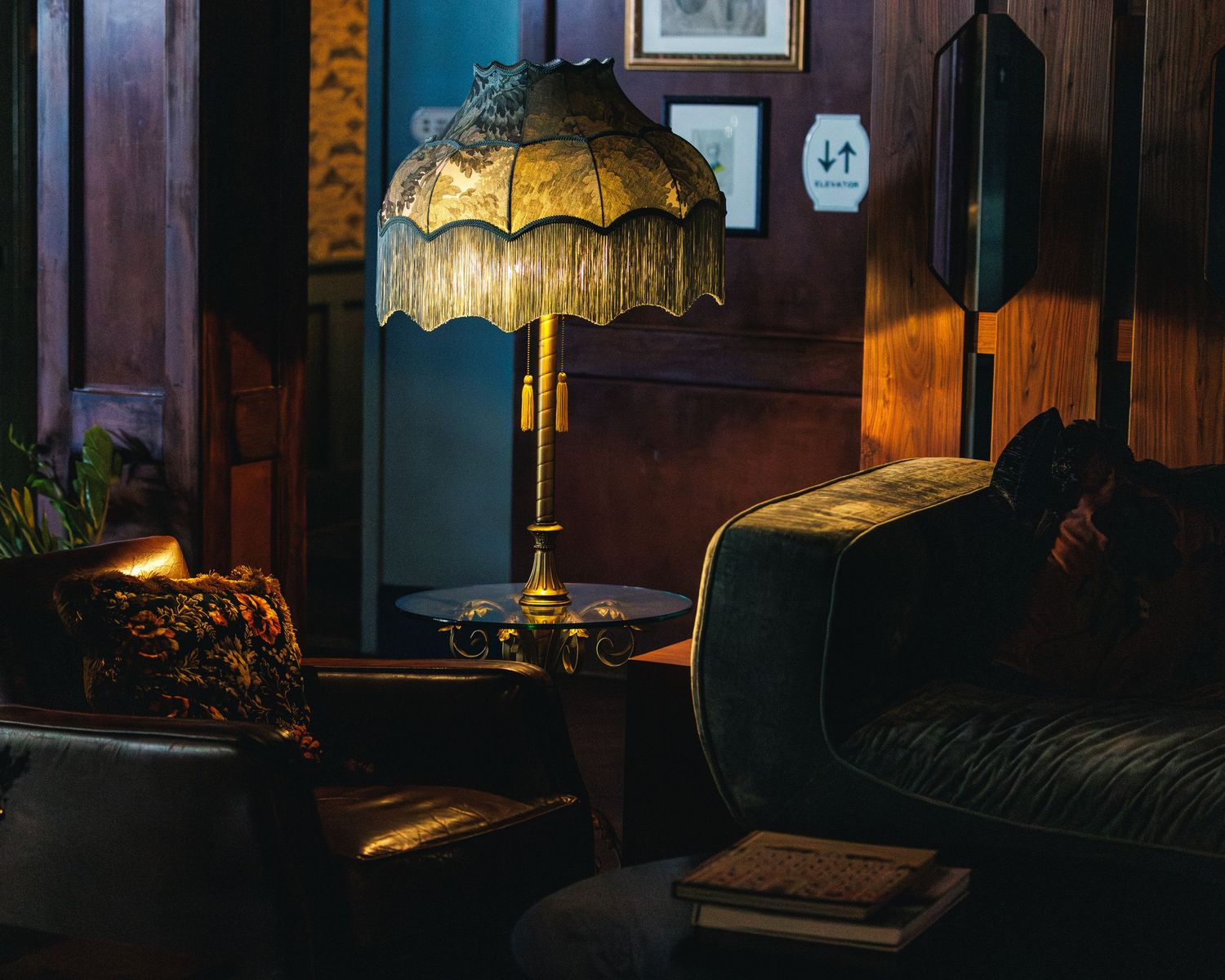 a dark moody interior shot showcasing a dark green lamp with fringes and a glass table. A leather chair with a patterned pillow and books