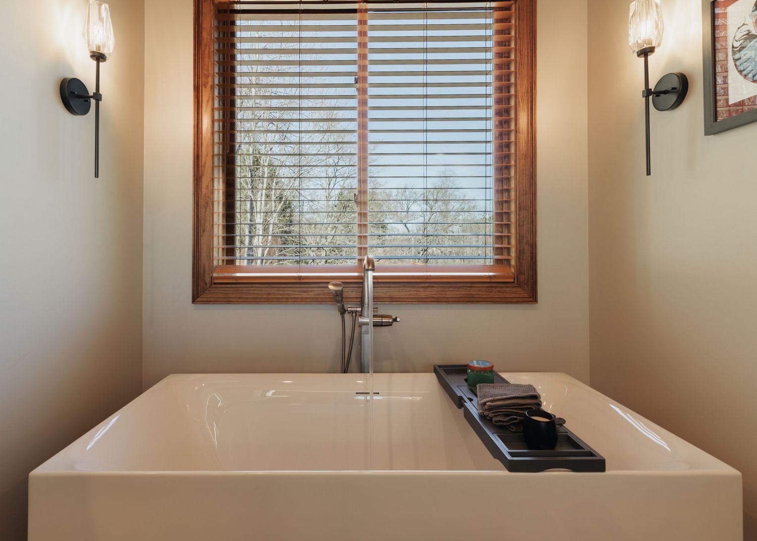 Large soaking bathtub surrounded by two wall sconces. A large window with wood shutters.