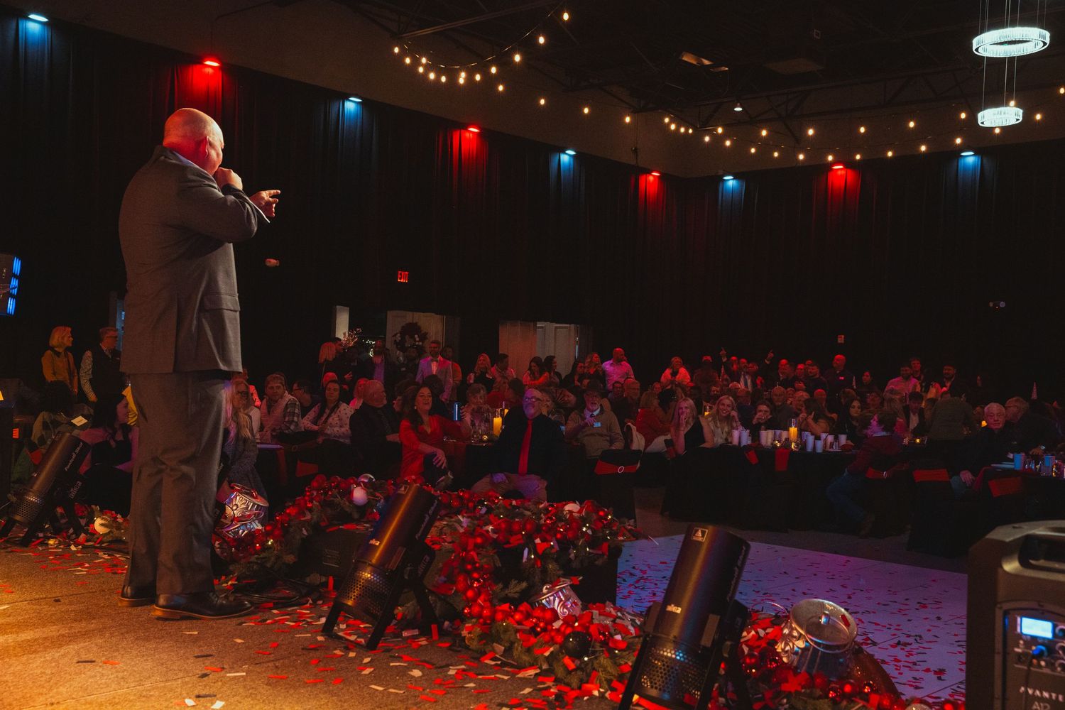 A man stands on stage and points out to the audience. The camera is facing the audience, only showing the man's back