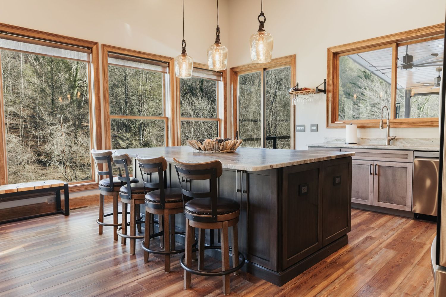 Large kitchen island with gray cabinets and three pendant lights hanging above it. Large windows surrounded by nature.