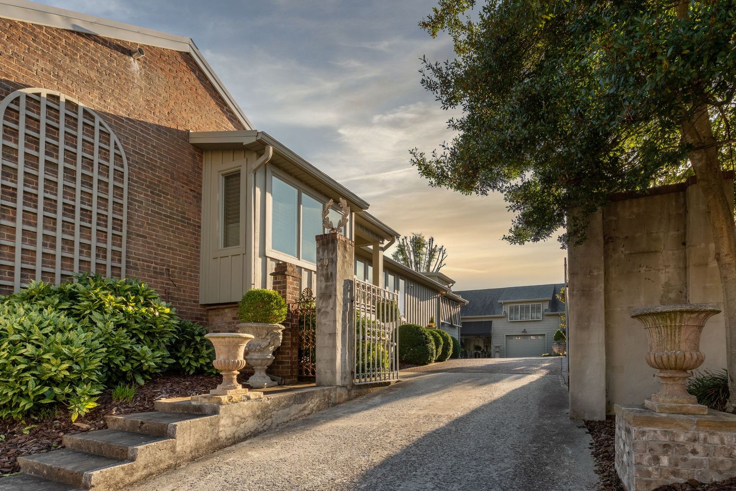 Exterior of home showing driveway entry. Sun beams are stretching across the facade. Green planters and bushes are lining the entry.