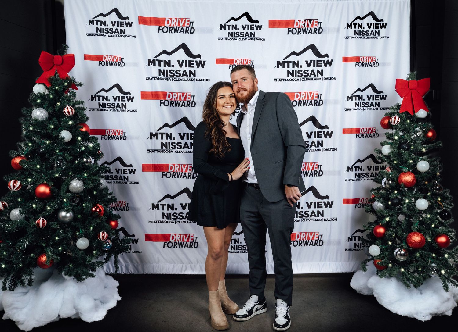 a man and a woman pose together surrounded by two Christmas trees with red bows on top of them