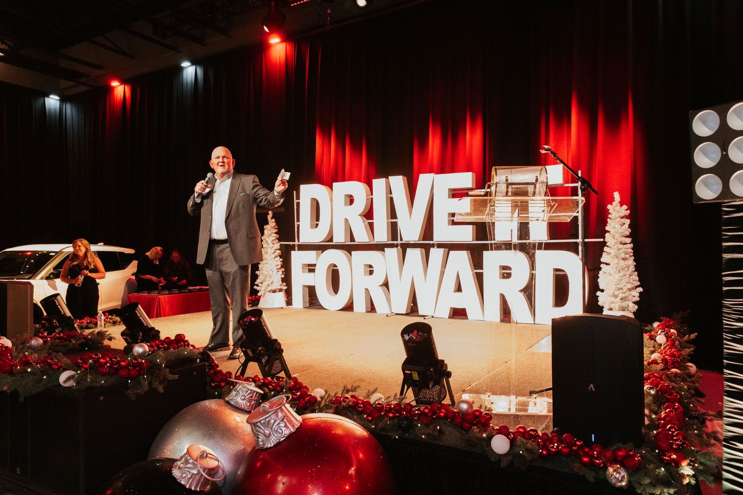A man stands on a backlit stage with the words Drive It Forward illuminated behind him. There are two large festive ornaments in front.