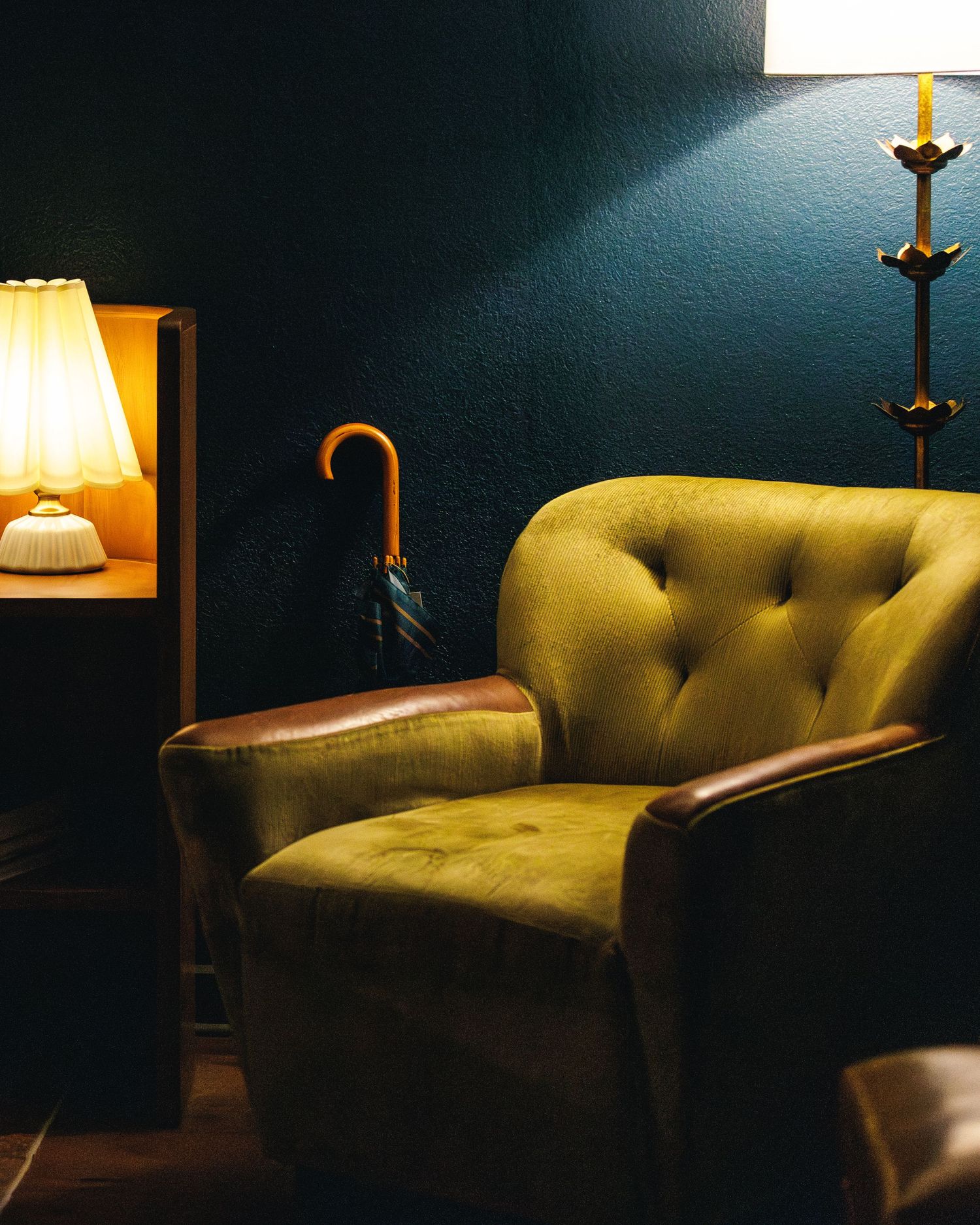 velvet sheen light green chair with a wooden handle umbrella resting against a dark, deep blue wall with lamps on either side