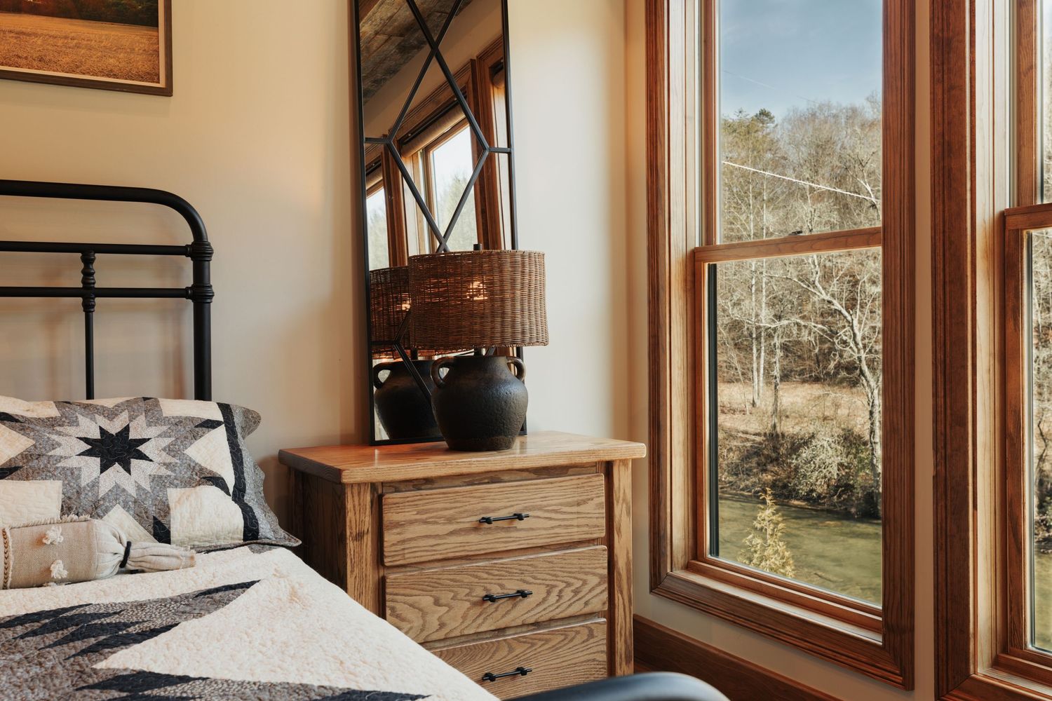 A bed with a white and blue star quilt on the left, a nightstand with a lamp in the center with large windows on the right showing a river