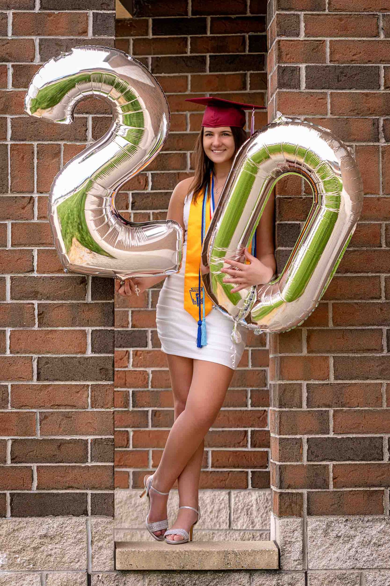 Graduate in white dress and red cap poses with large silver 