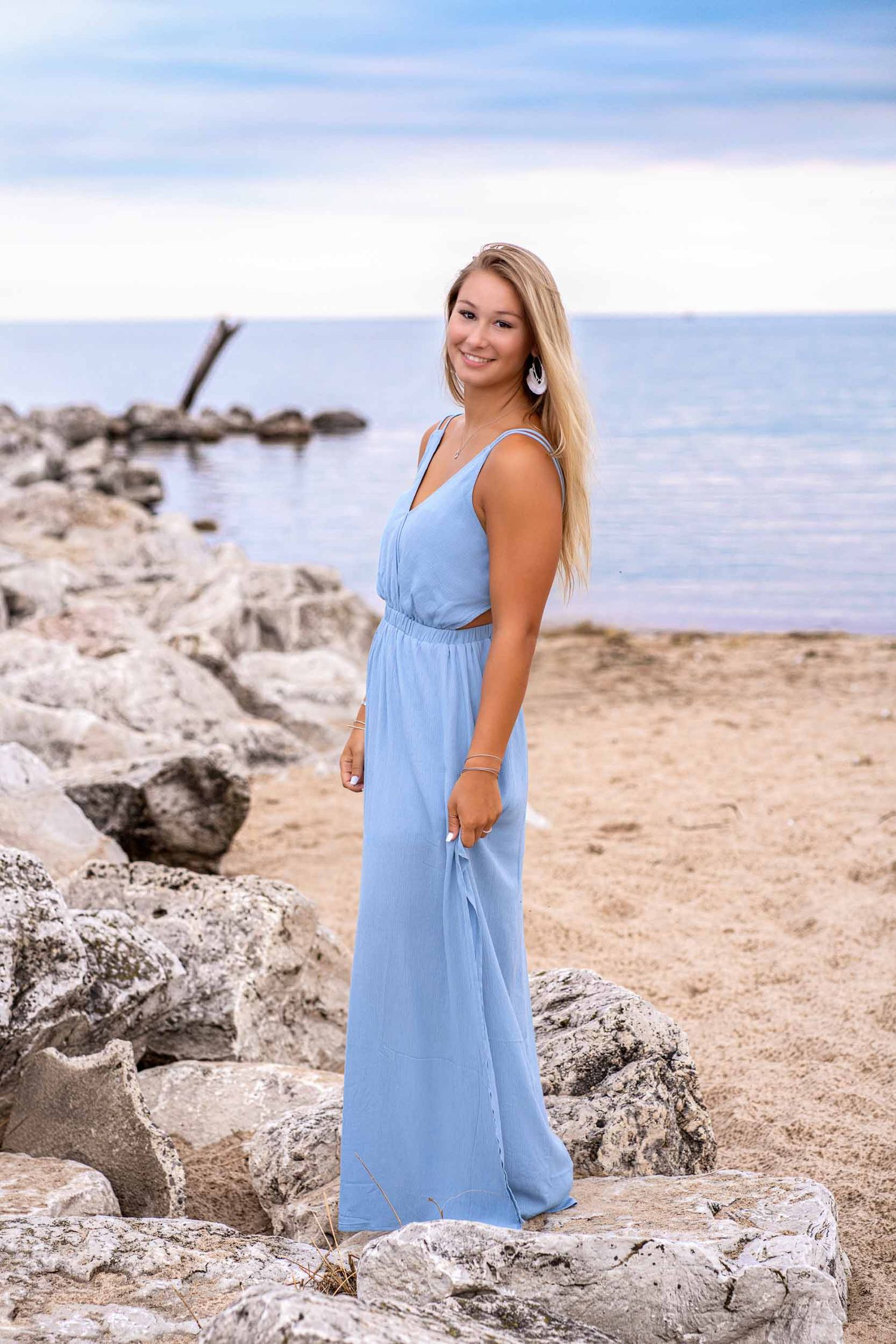 Woman in light blue summer dress poses on rocky beach with ocean in background.
