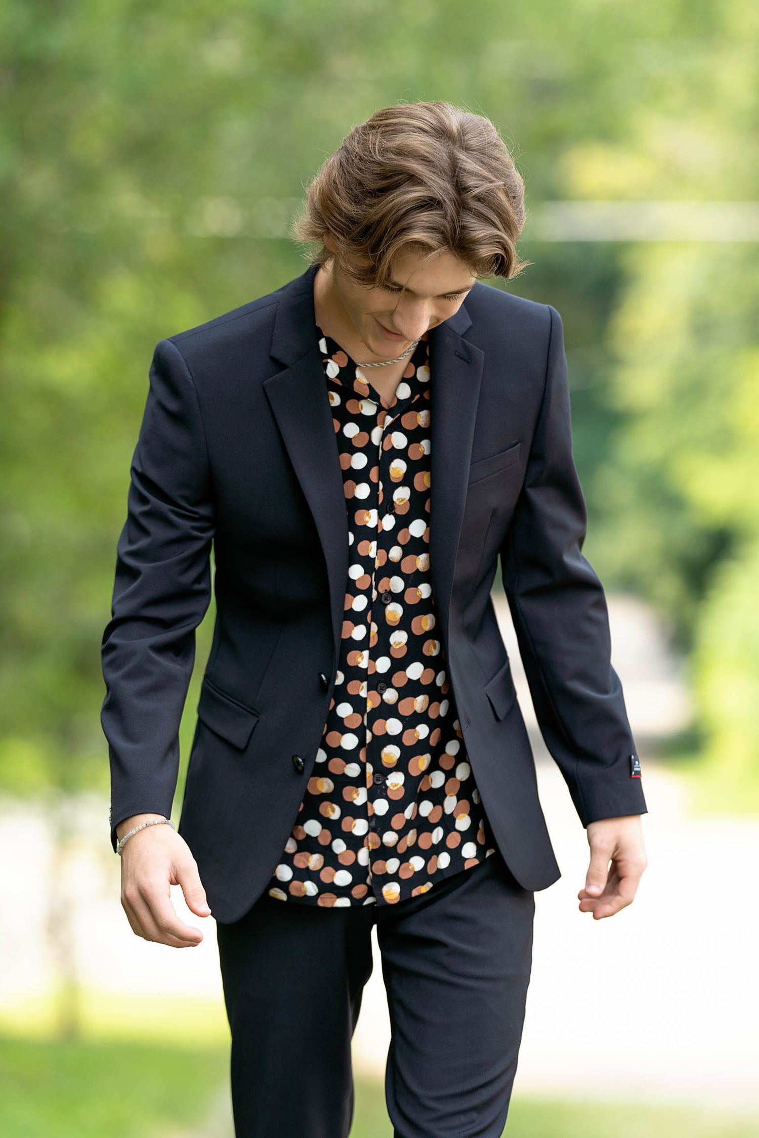 A person wearing a navy suit with a patterned button-up shirt poses outdoors against a blurred green background.