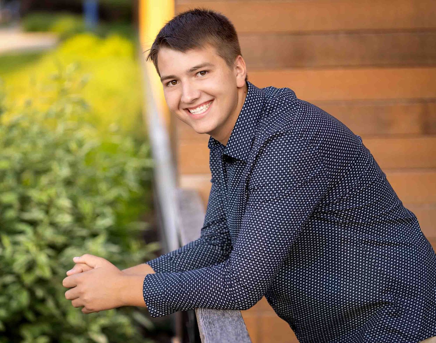 A person wearing a navy polka dot shirt sits casually on outdoor steps while smiling at the camera.