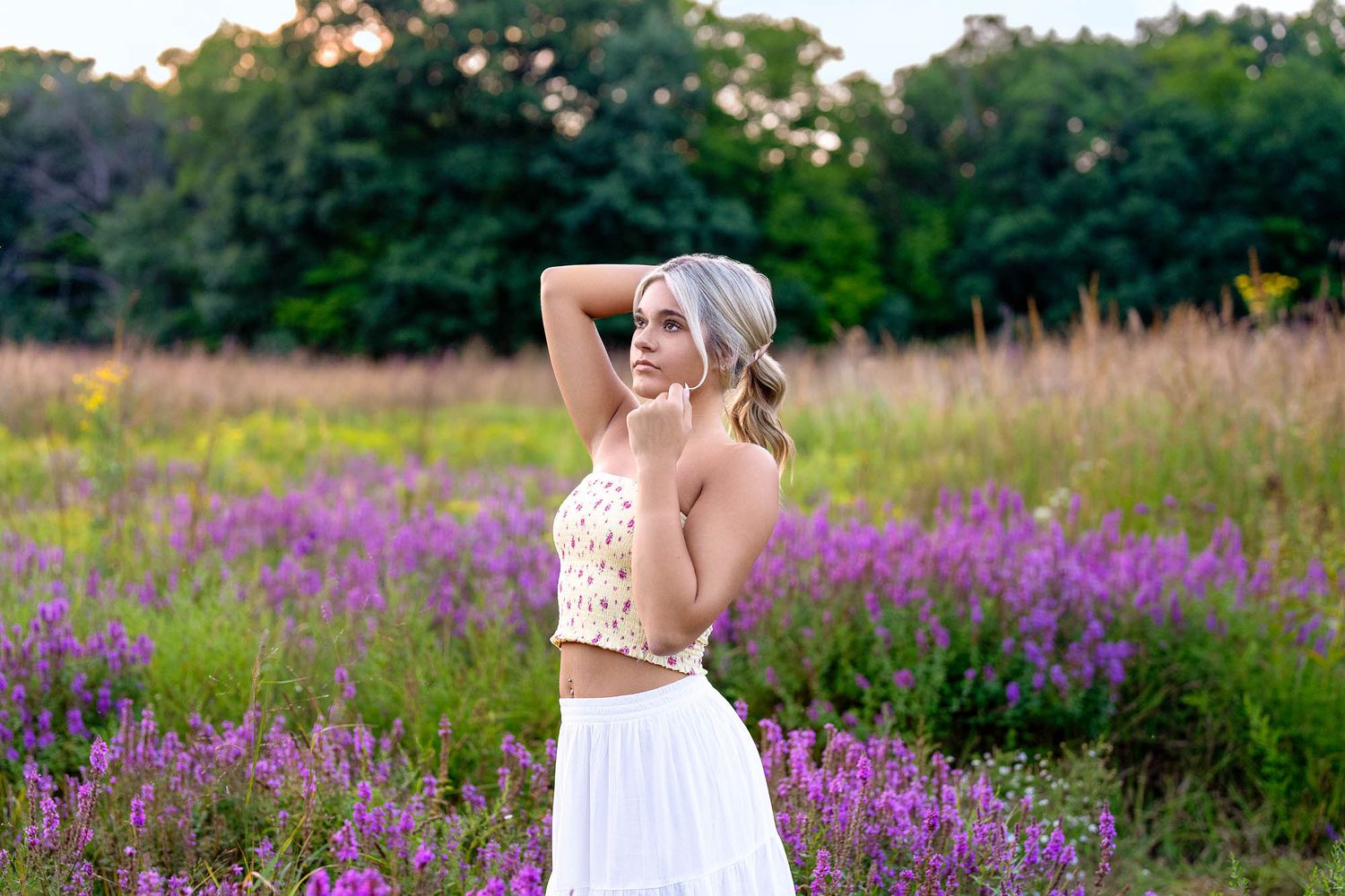 Blonde woman in floral crop top and white skirt posing in a vibrant purple wildflower field at sunset.