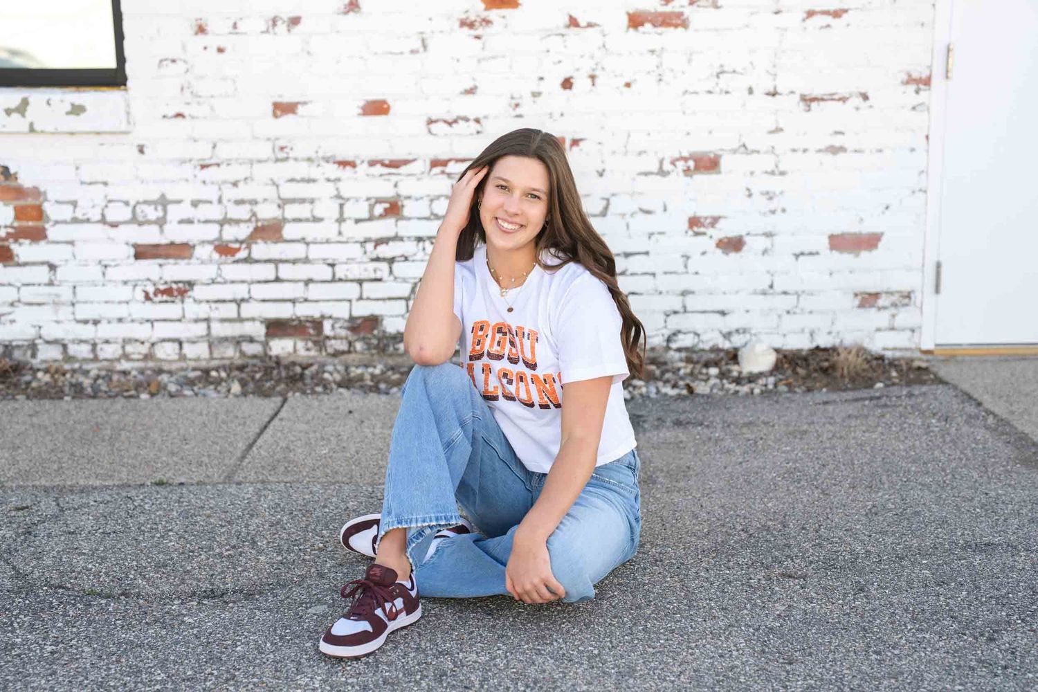 Student in casual BGSU spirit wear sits cross-legged on ground against white brick wall background.
