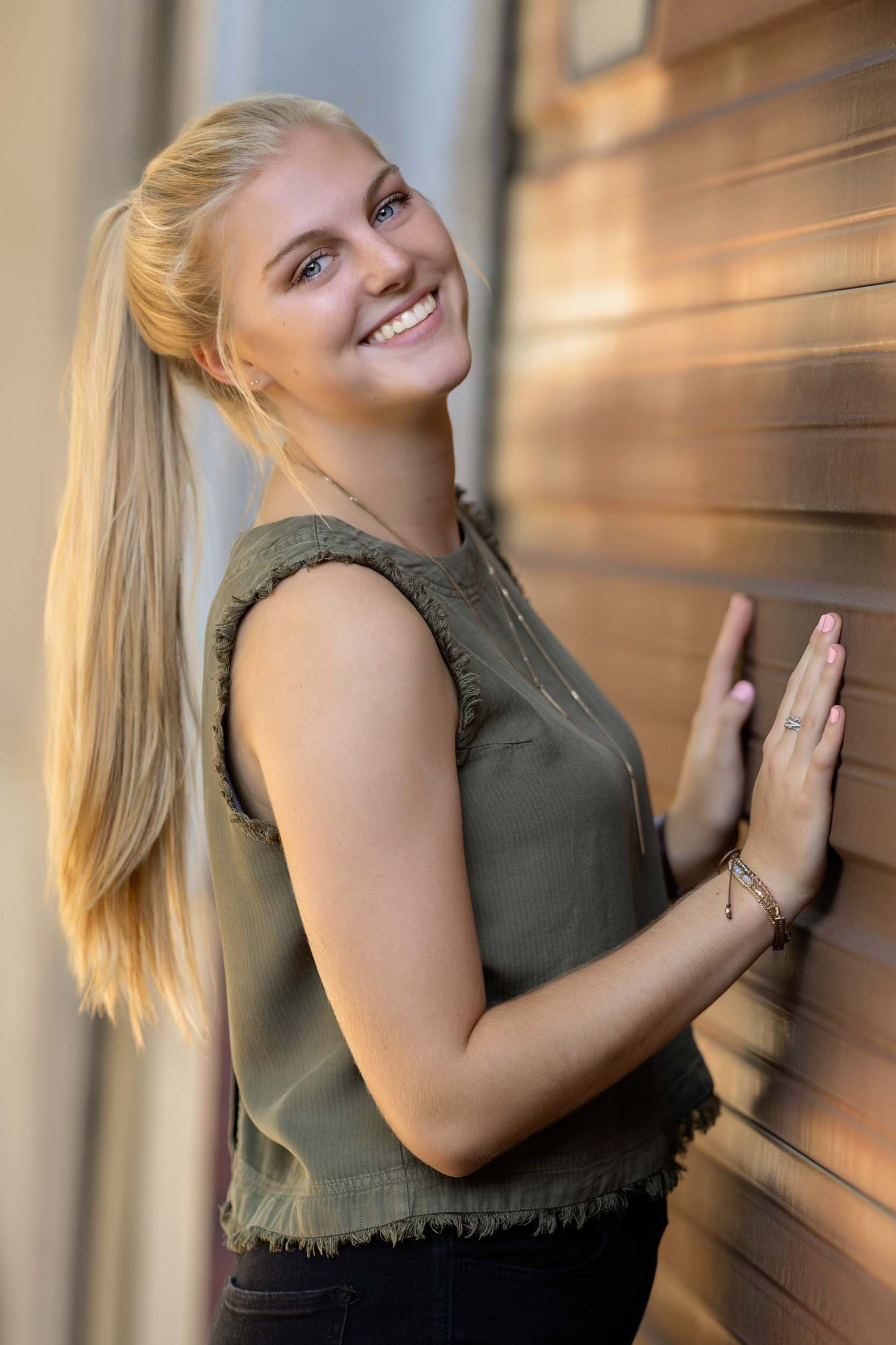 Woman in olive green sleeveless top poses against wooden wall in natural lighting.