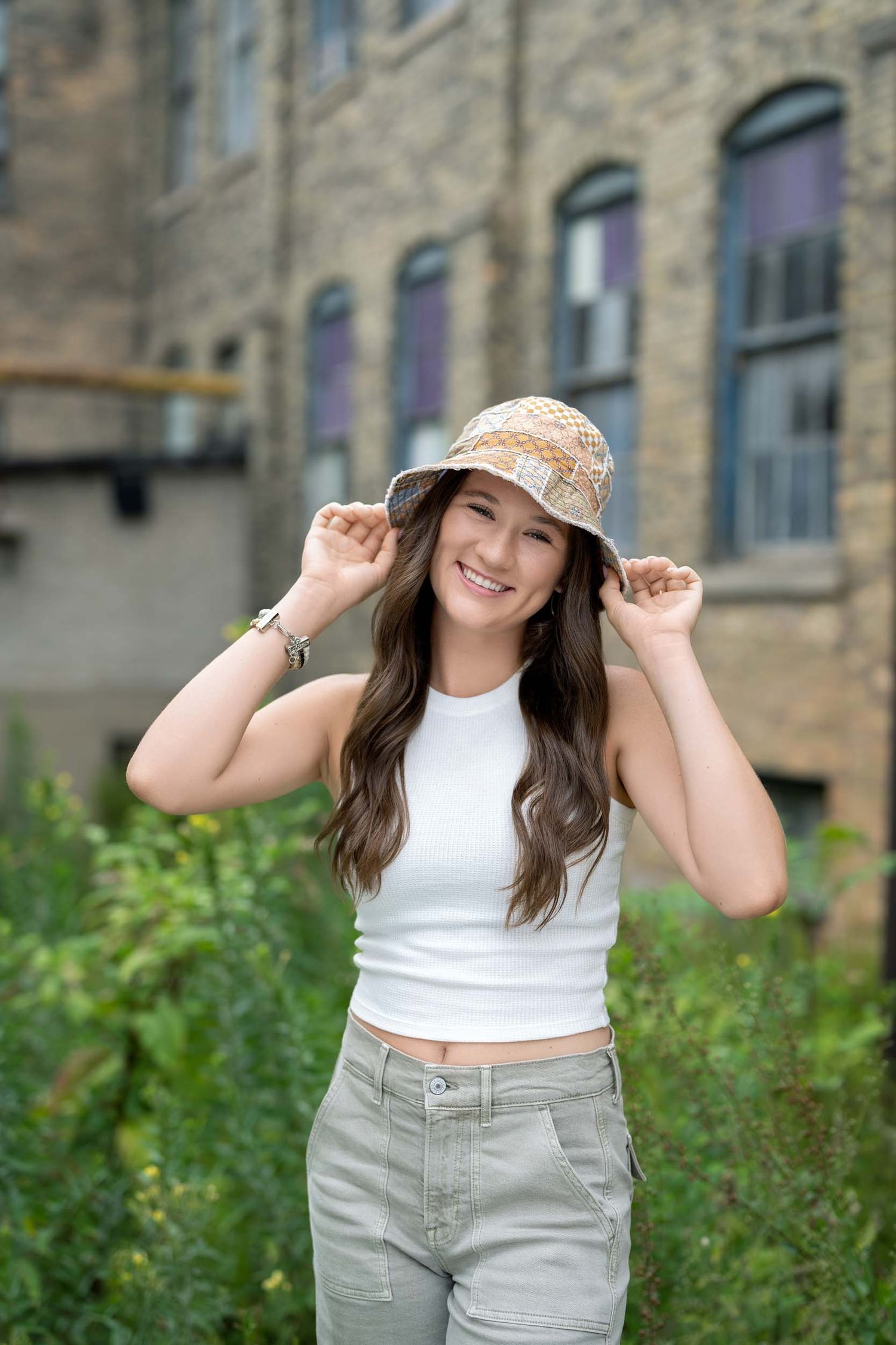 A person wearing a white crop top and khaki pants poses playfully in front of a stone building.