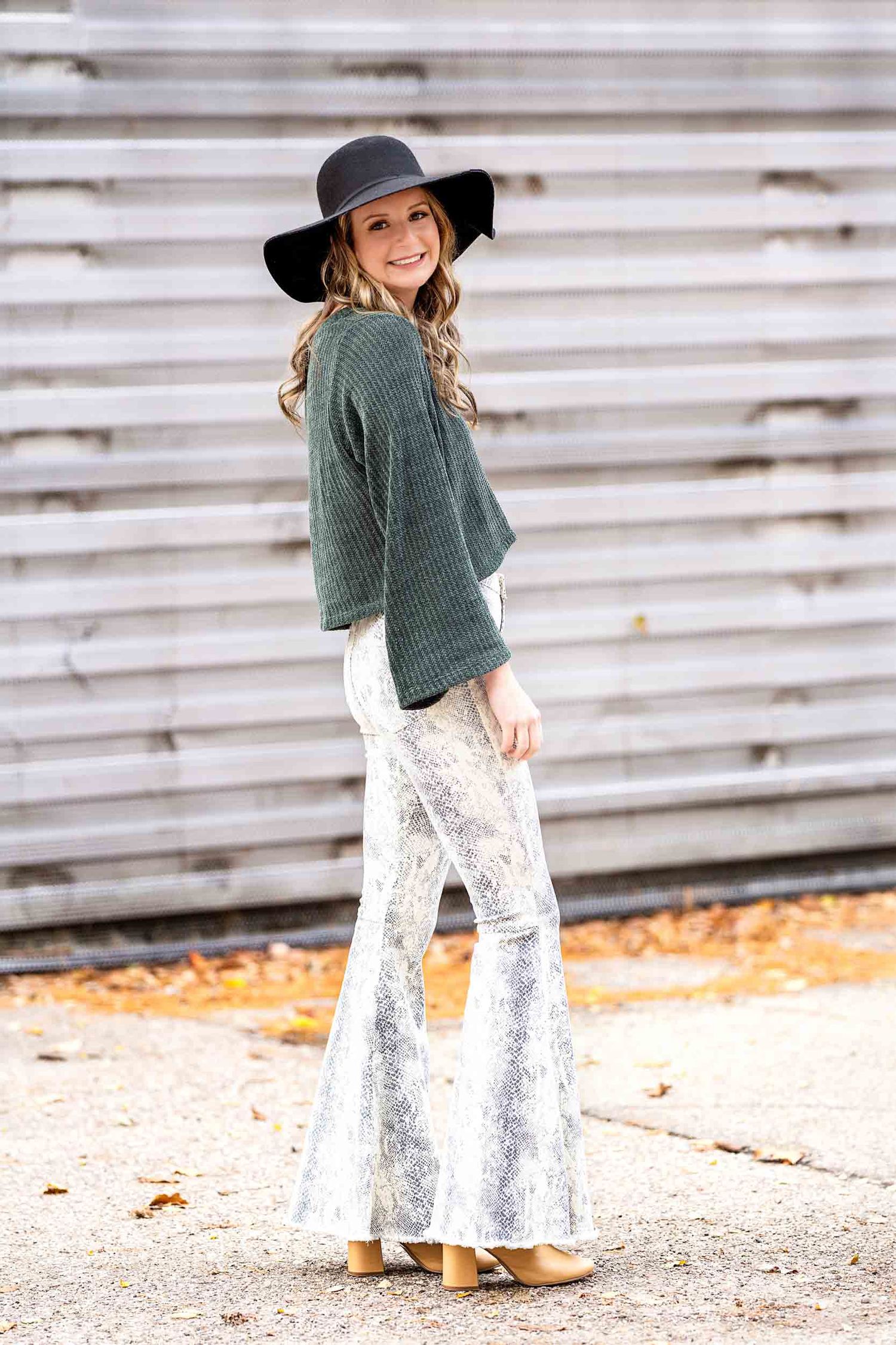 Stylish fall outfit featuring white snakeskin flare pants, dark sweater and wide brim hat with autumn leaves in background.