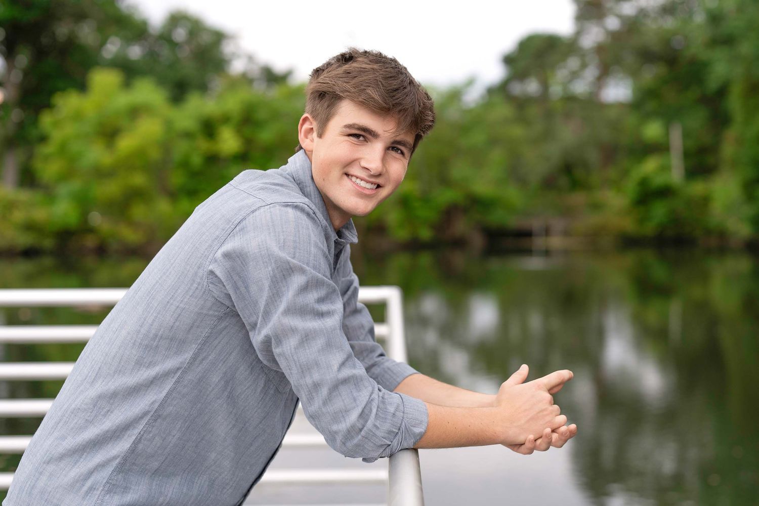 A person in a gray shirt stands near a white fence by a pond on a sunny day.