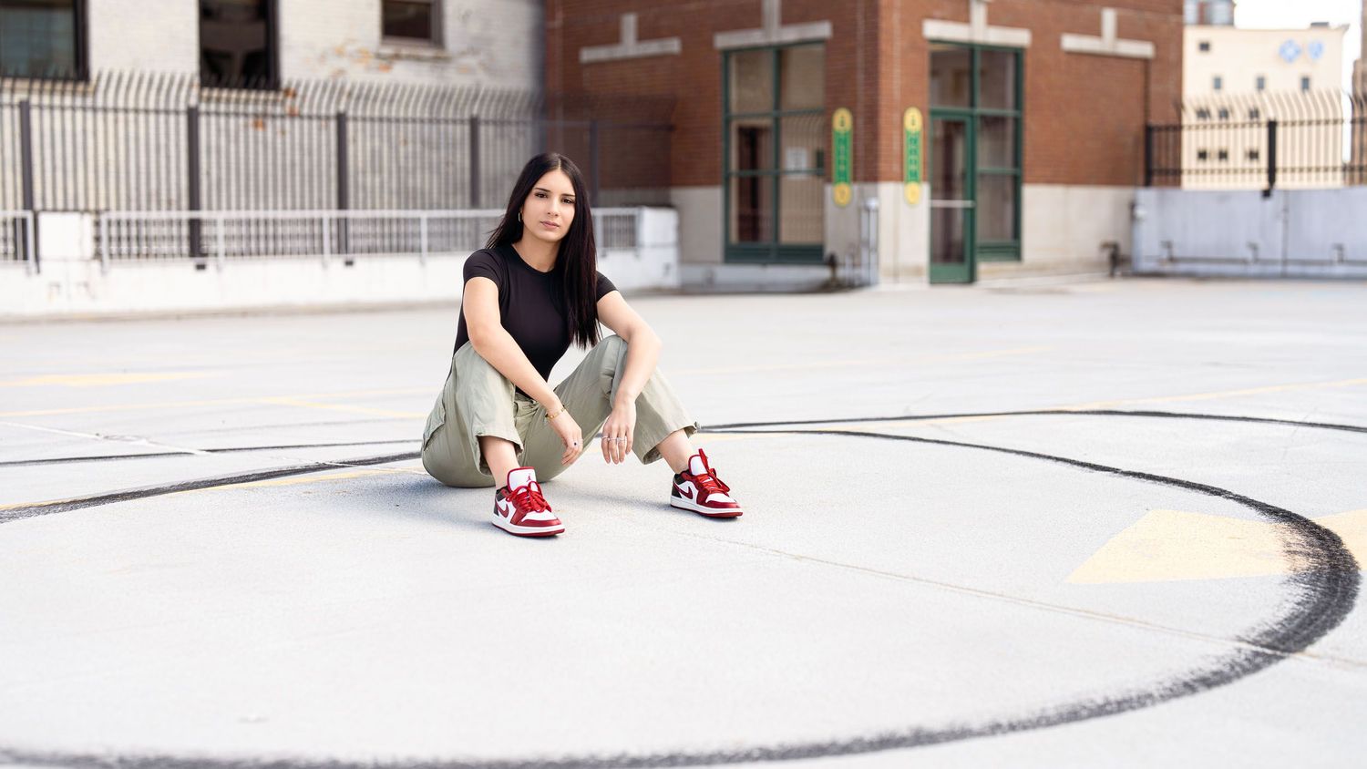 A young woman sits cross-legged on an outdoor court wearing cargo pants and red sneakers, with a brick building behind her.