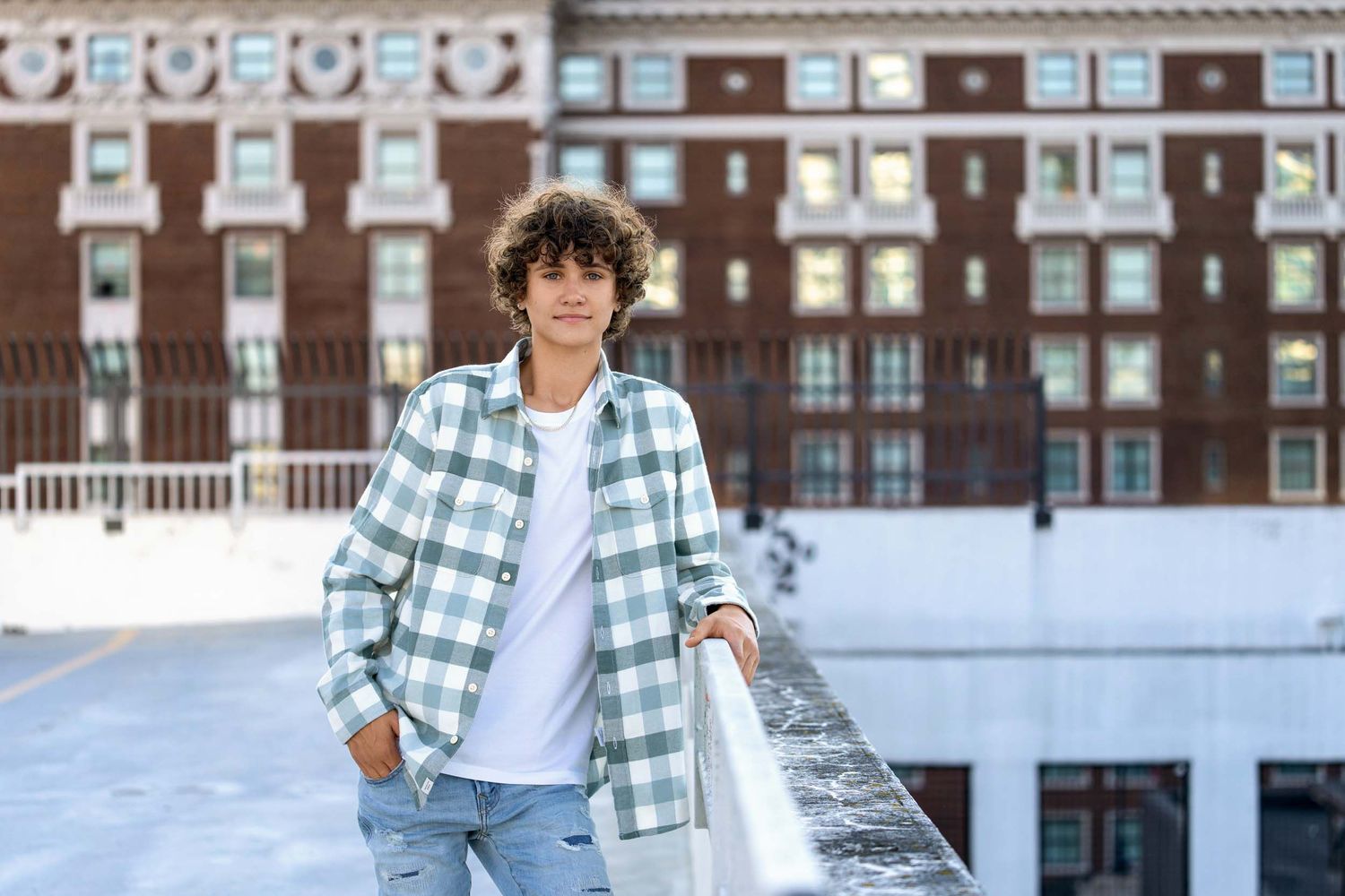 A person wearing a plaid shirt and white t-shirt poses in front of a brick building.