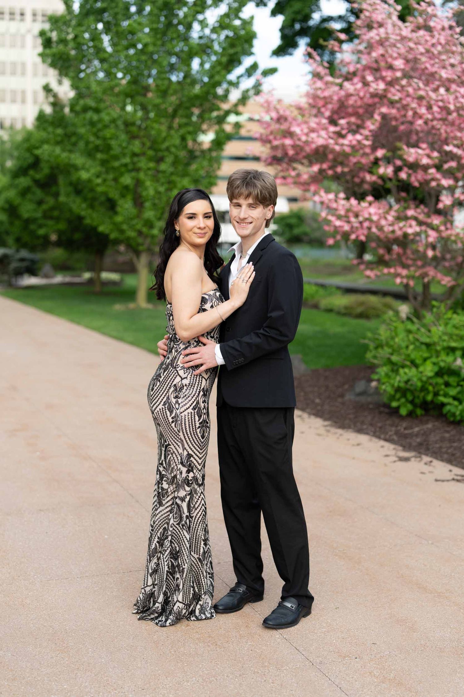 A formal couple poses outdoors on a pathway with blooming cherry trees and greenery in the background.